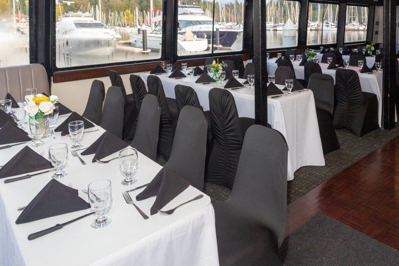 Interior of a dining area on a boat with tables set for a formal event. White tablecloths, black chair covers, and black napkins are visible. Outside, there are boats and a marina in the background.