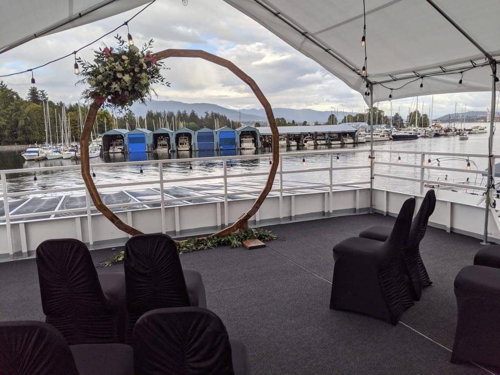 Outdoor wedding setup on a boat with black covered chairs, a large circular wooden arch adorned with flowers, overlooking a marina with boats and covered docks.