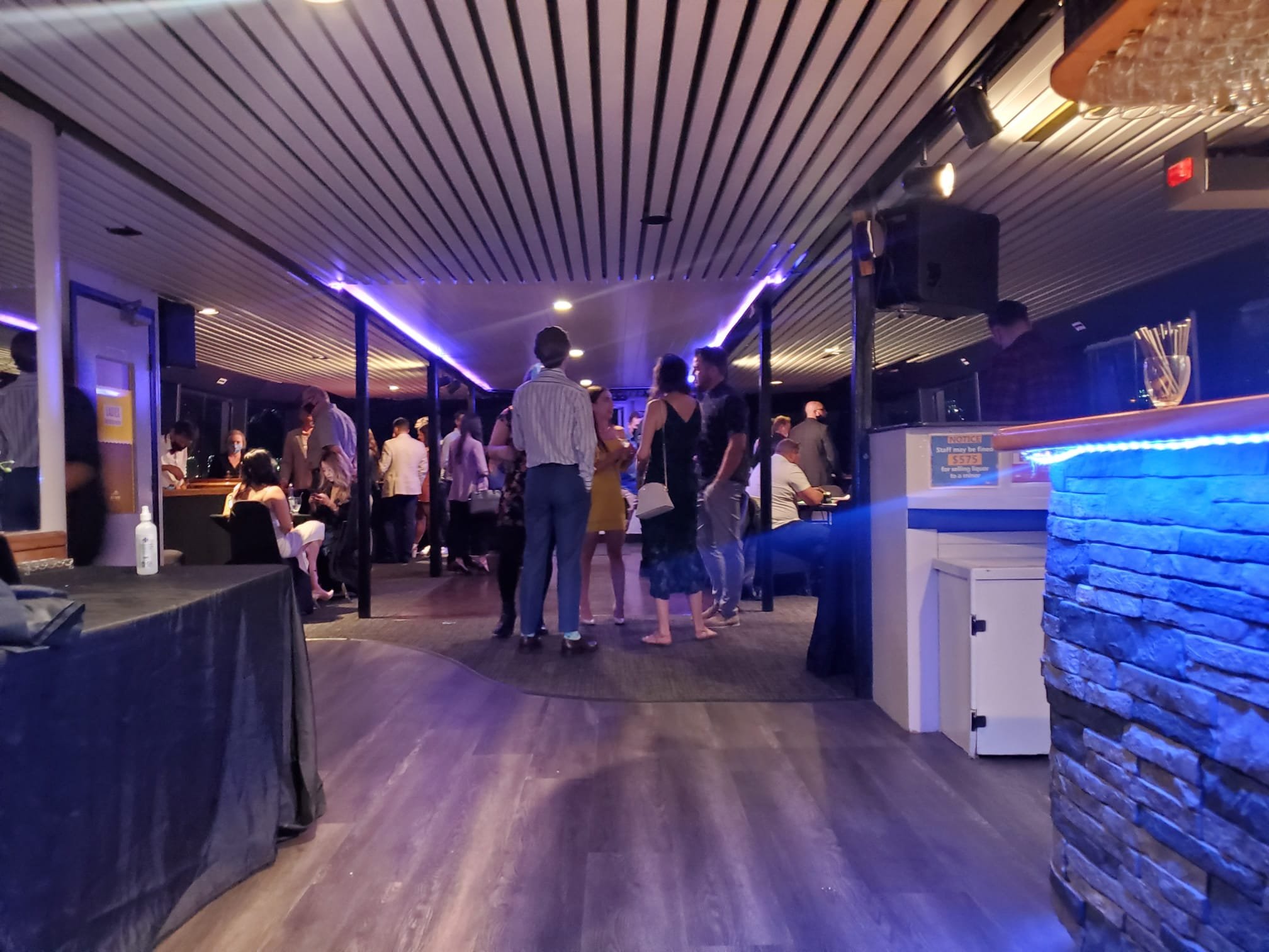 Indoor lounge with people socializing, wood flooring, purple ambient lighting, stone accent wall, tables and chairs, and a bar area.