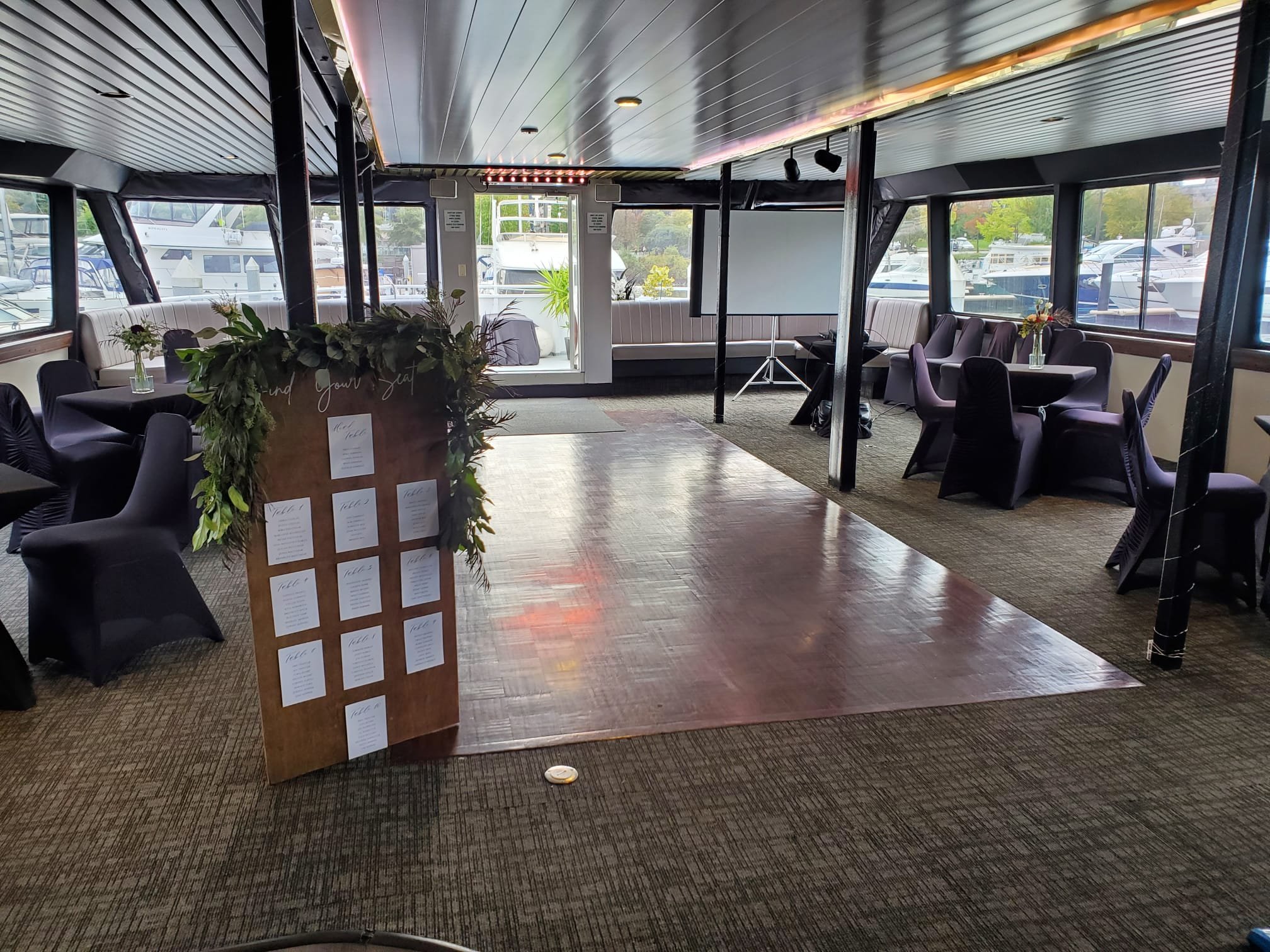 Indoor event venue on a boat with tables and chairs, covered in dark fabric, surrounding a wooden dance floor. A seating chart with plants atop is displayed near the entrance. Large windows show a view of moored boats outside.