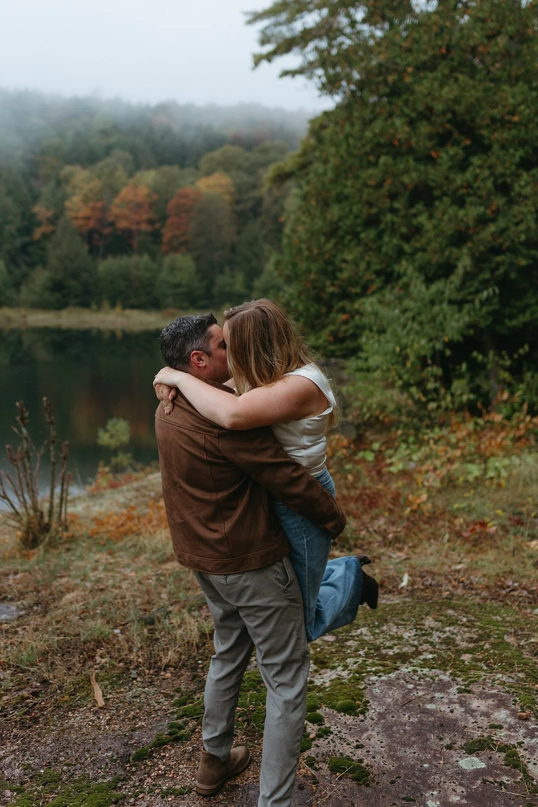A man lifting and kissing a woman outdoors near a lake with trees in fall foliage.