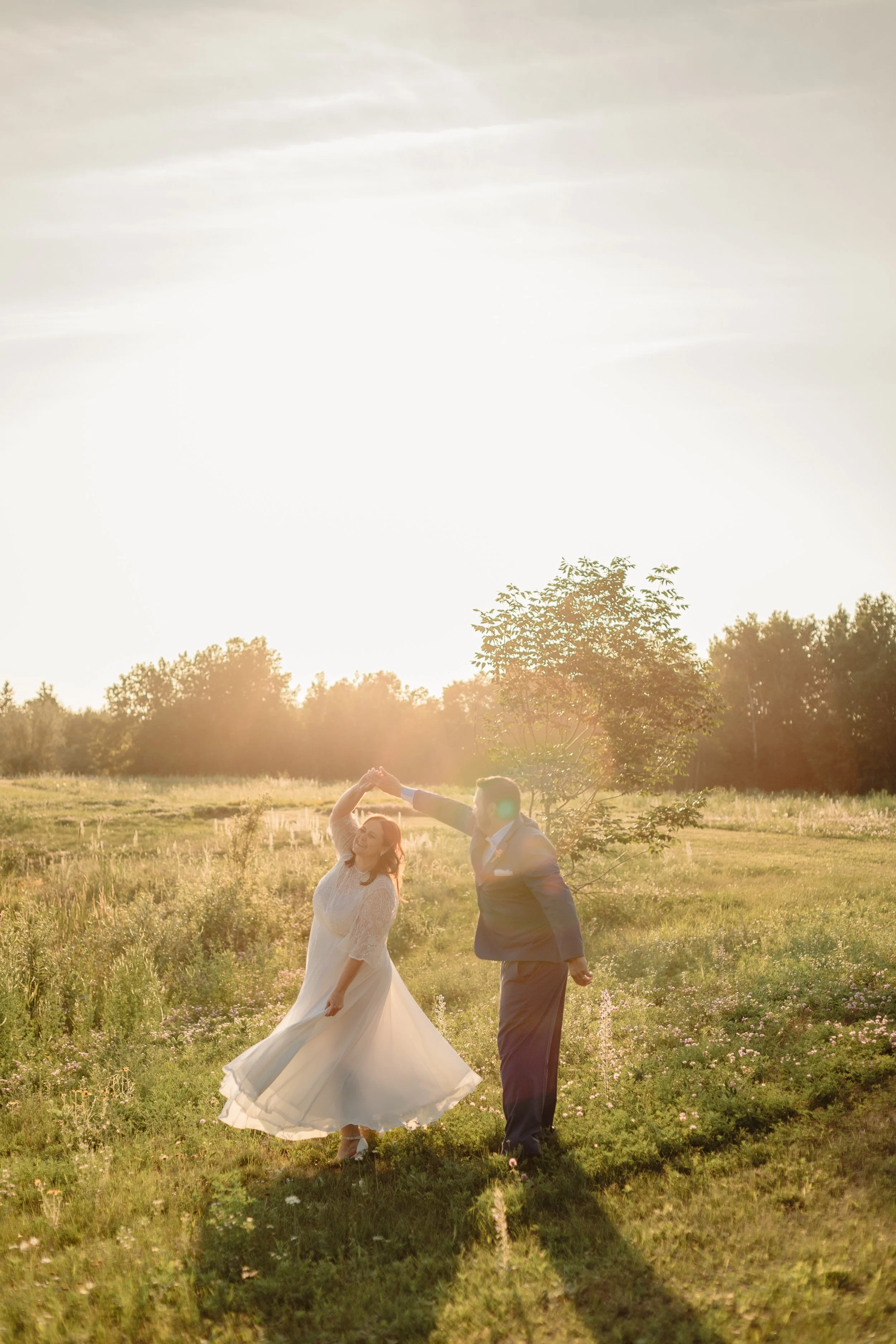 A bride and groom dancing in a sunlit field during sunset, with the bride wearing a white dress and the groom in a blue suit, surrounded by greenery.