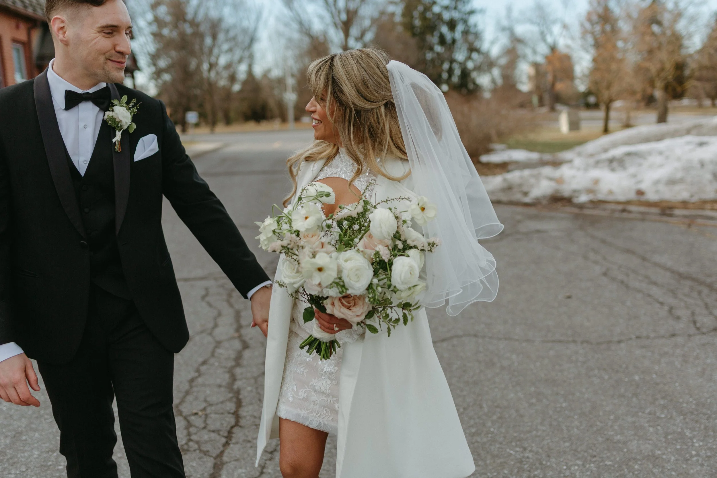 A bride and groom holding hands outdoors on a neighborhood street during winter or early spring, with trees and snow patches in the background.