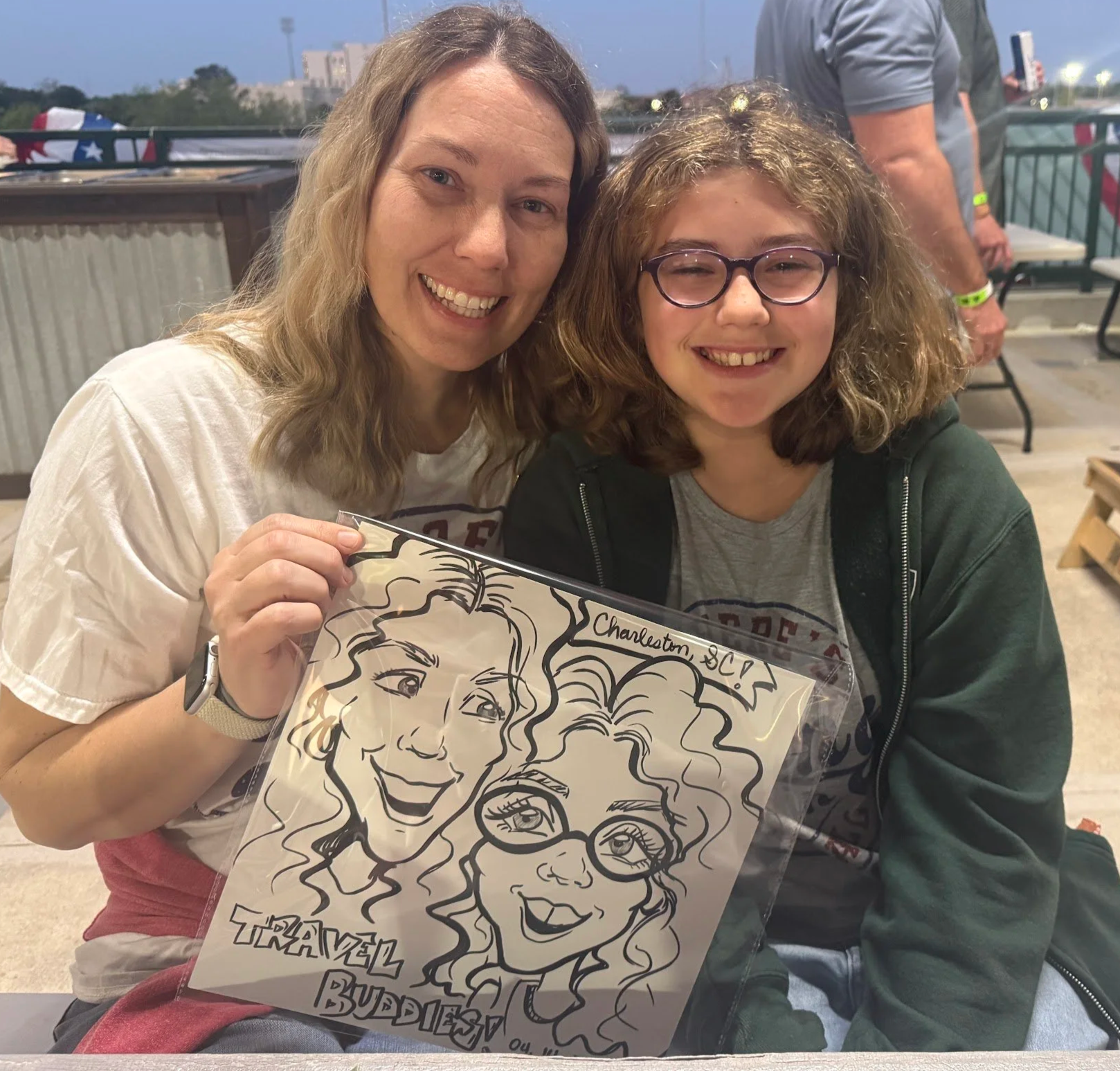 Two women smiling and holding a caricature drawing of themselves at an event. The woman on the left has long wavy hair, is wearing a white shirt, and has an Apple Watch on her left wrist. The woman on the right has curly hair, glasses, and is wearing a gray T-shirt under a dark zip-up hoodie. The caricature drawing features exaggerated facial features of both women and includes the text "Traditional Birthday" and "Charleston" on it. The background shows an outdoor or semi-outdoor setting with a few people and tables.