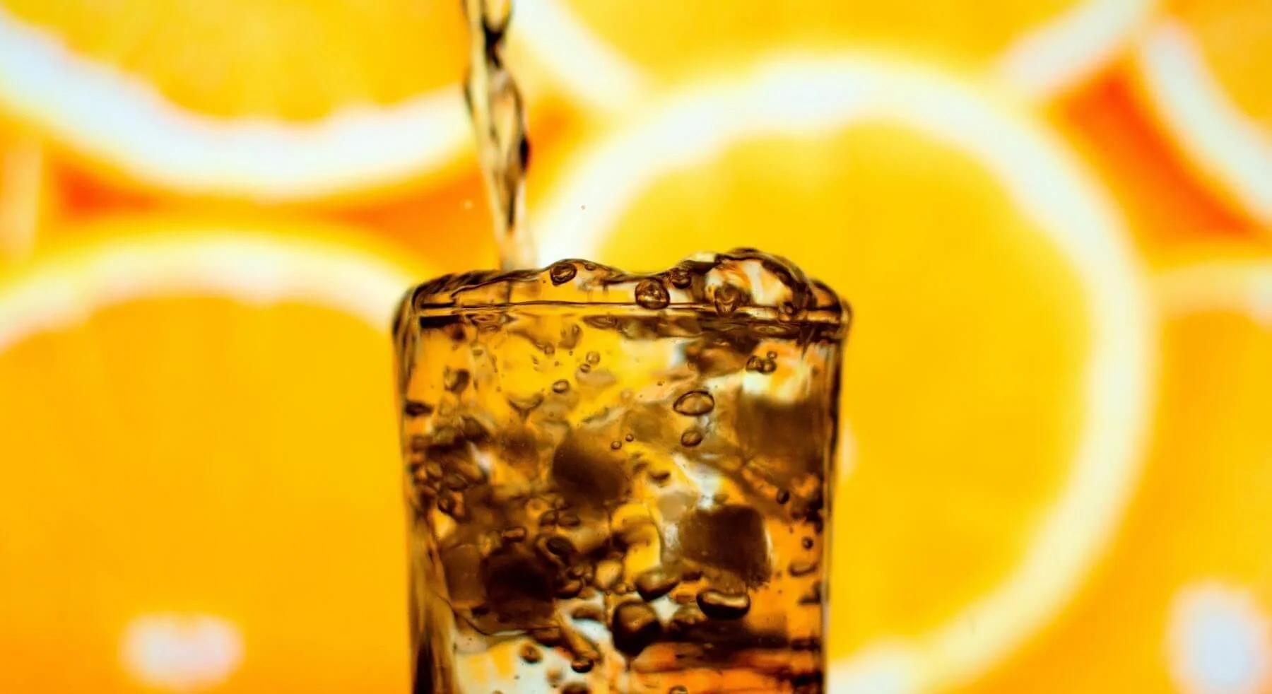 A close-up of a glass with ice cubes, orange juice is being poured into it, with whole oranges in the background.
