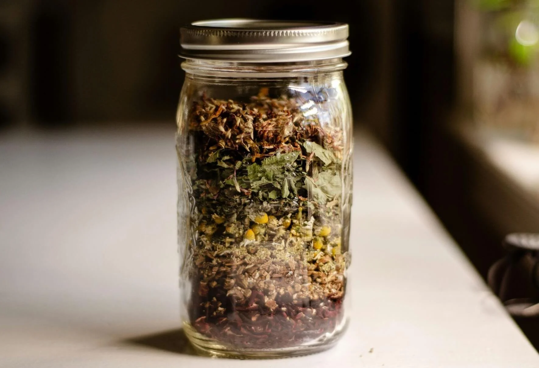 A glass jar filled with assorted dried herbs, flowers, and botanicals on a white surface.