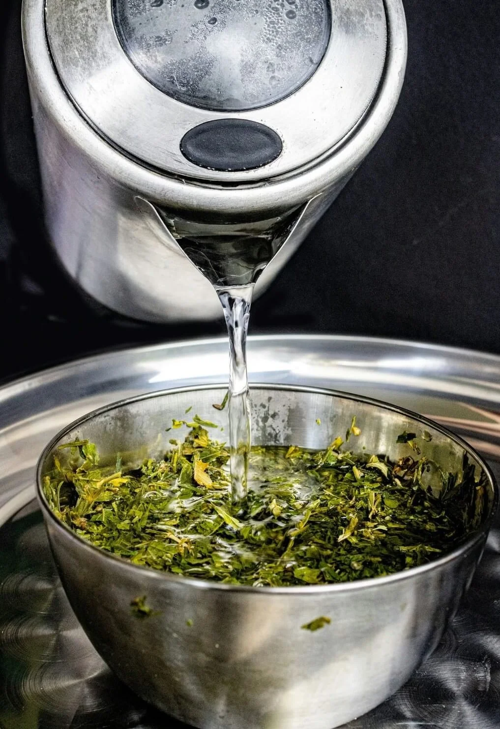 Hot water being poured from a stainless steel kettle into a bowl of fresh green herbs.