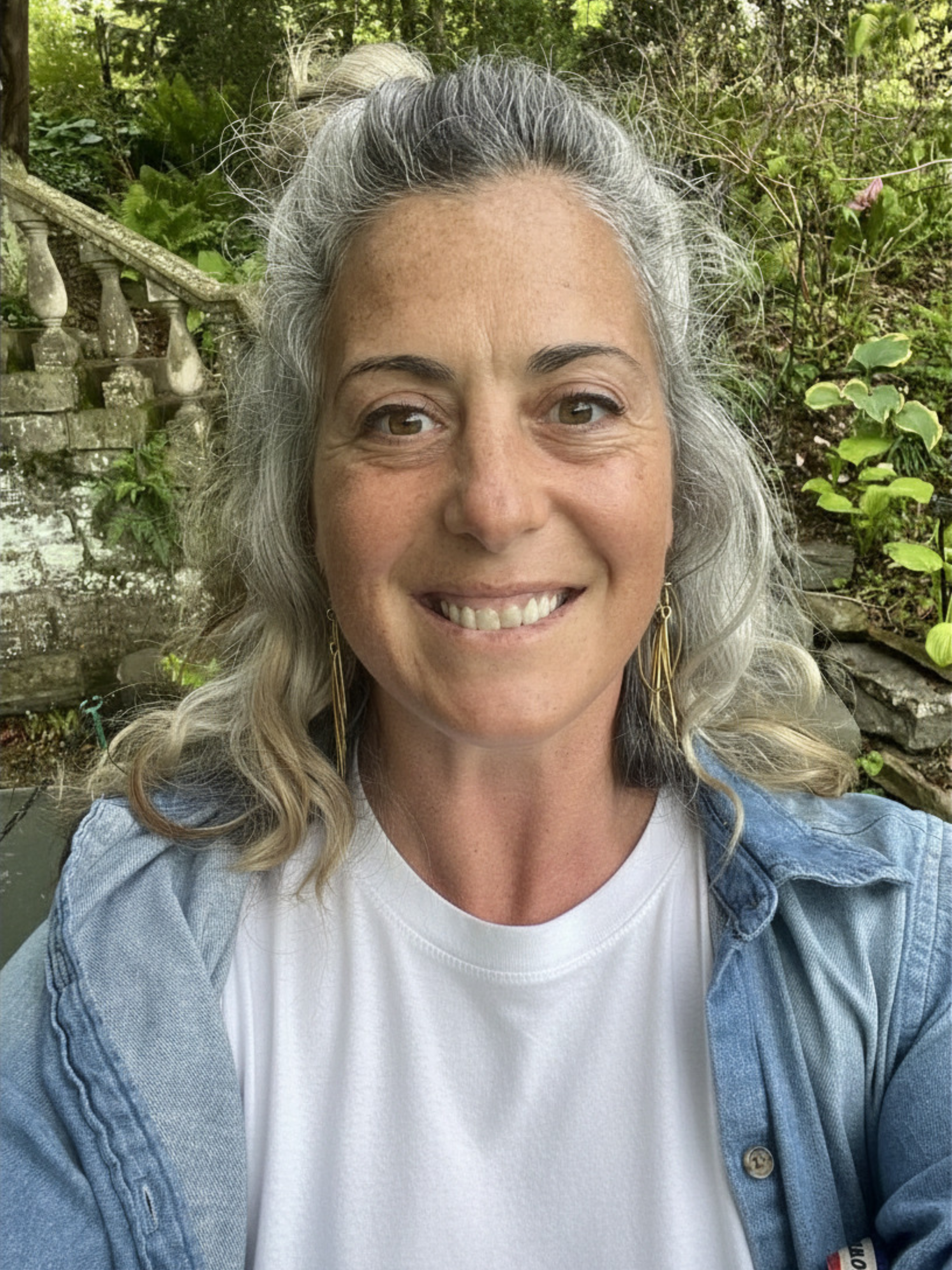 A woman with curly brown hair wearing a pink sweater and blue jeans, smiling and standing indoors near a rustic wood wall with white paint accents.