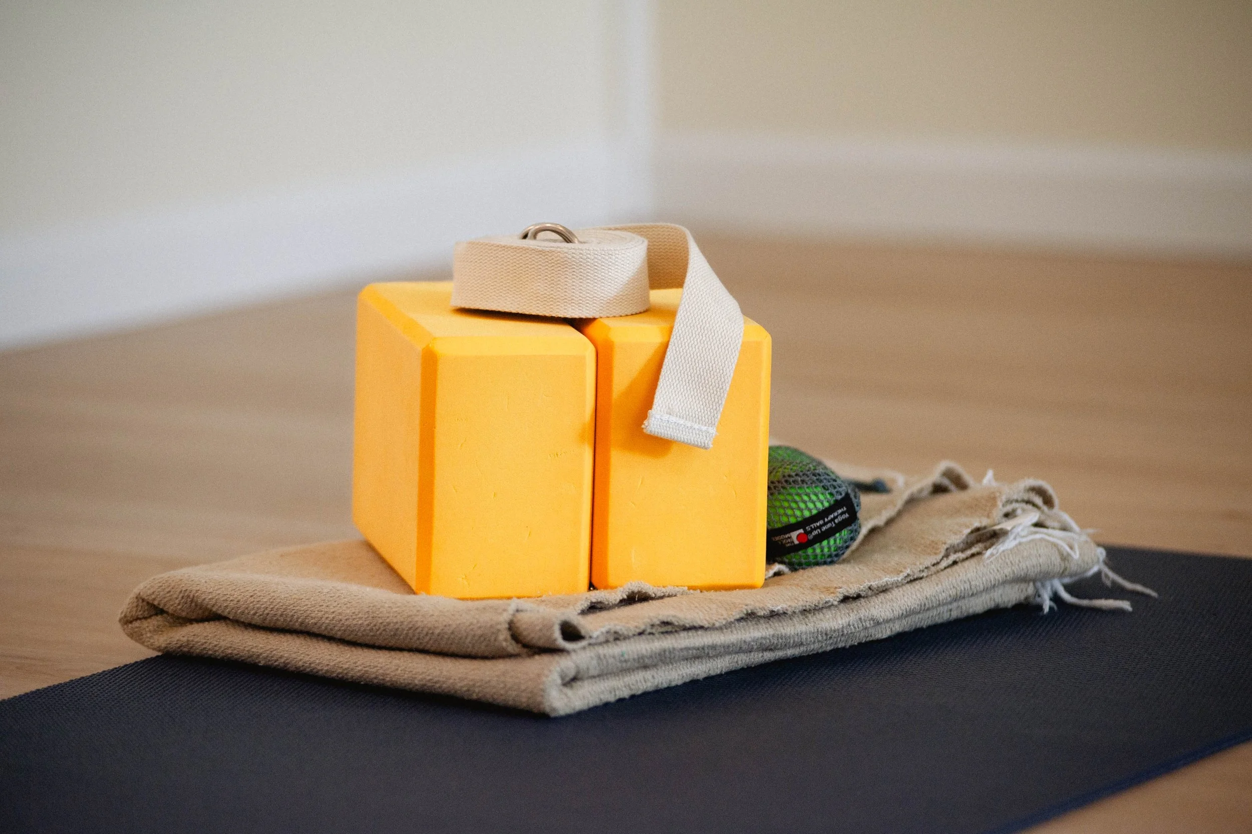 A yellow yoga block, a folded beige towel, a green and black massage ball, and a beige strap are arranged on a black yoga mat on a wooden floor.