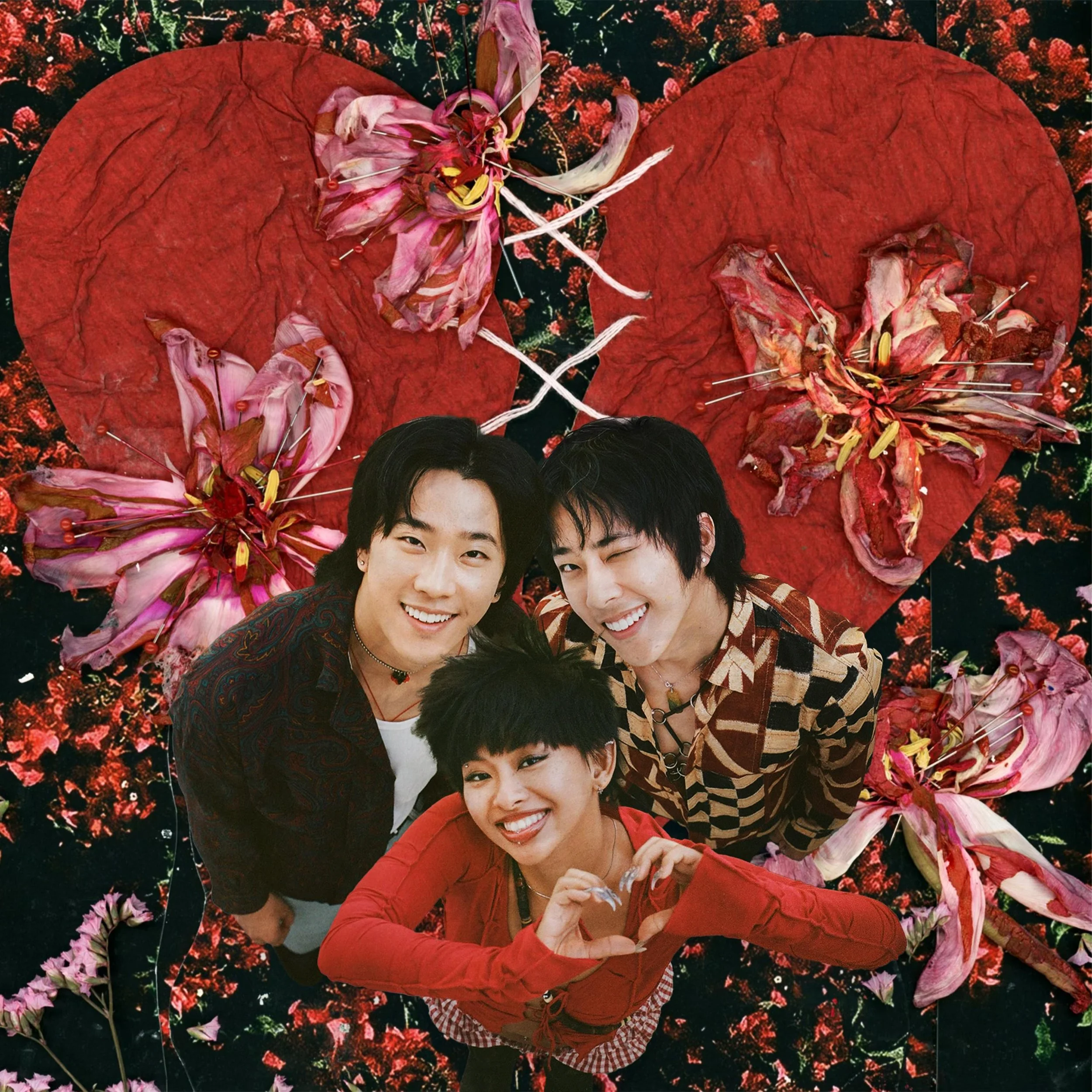 Three young people smiling and posing for a photo against a background of a large red heart and pink flowers, with some flower petals pinned with sewing needles.