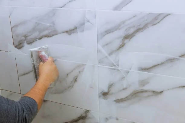 Person using a trowel to apply white marble-patterned tiles on a wall.