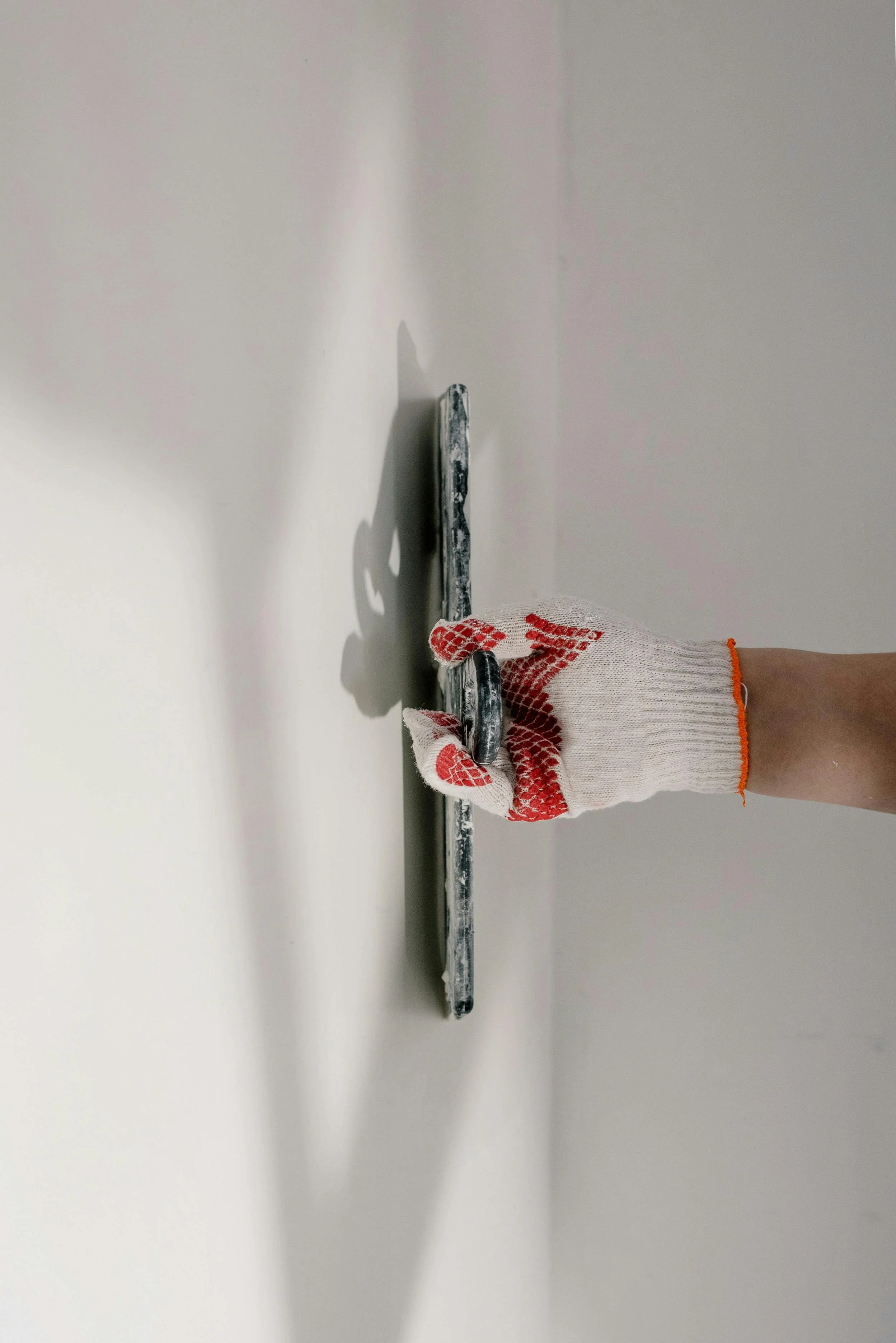 A person wearing a white glove with red dots, holding a metal scraper against a white wall during painting or drywall work.