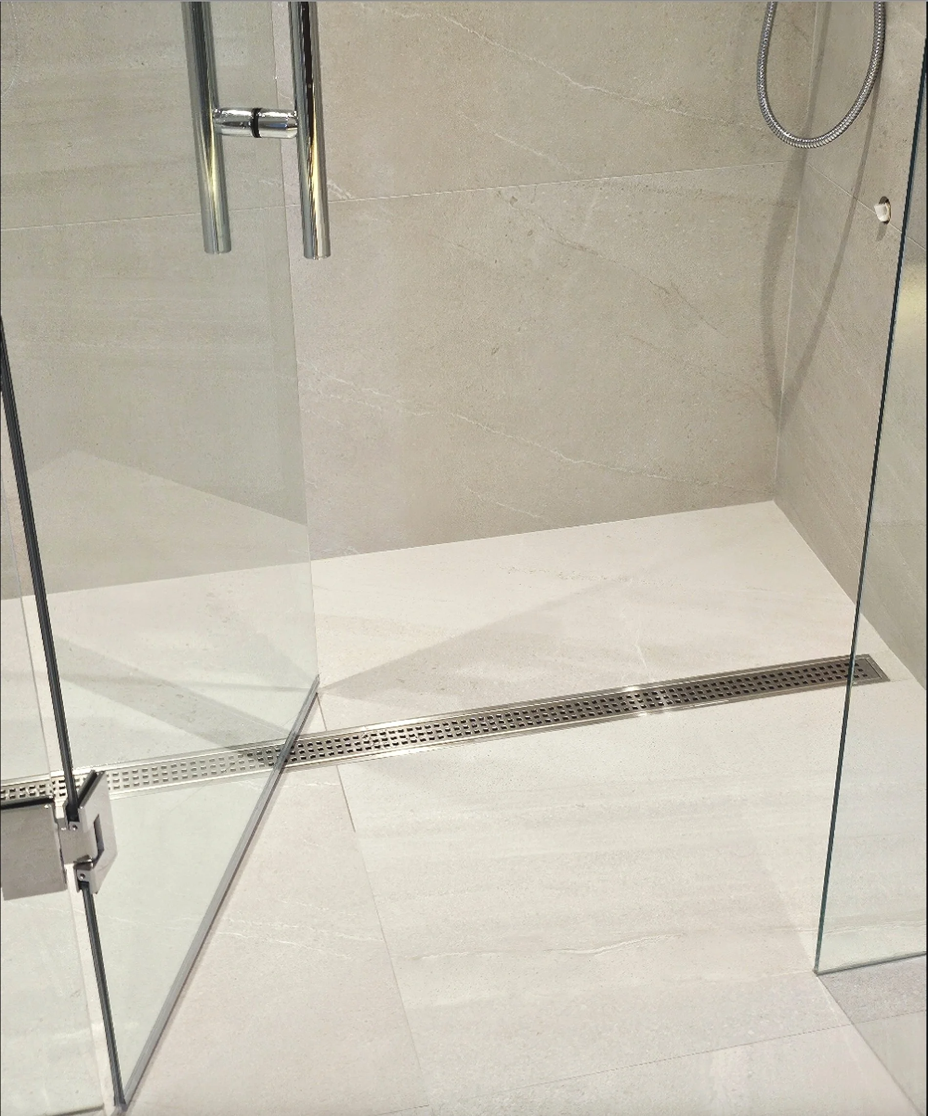 Modern walk-in shower with glass doors, beige wall tiles, and a metal drain on the light-colored tiled floor.