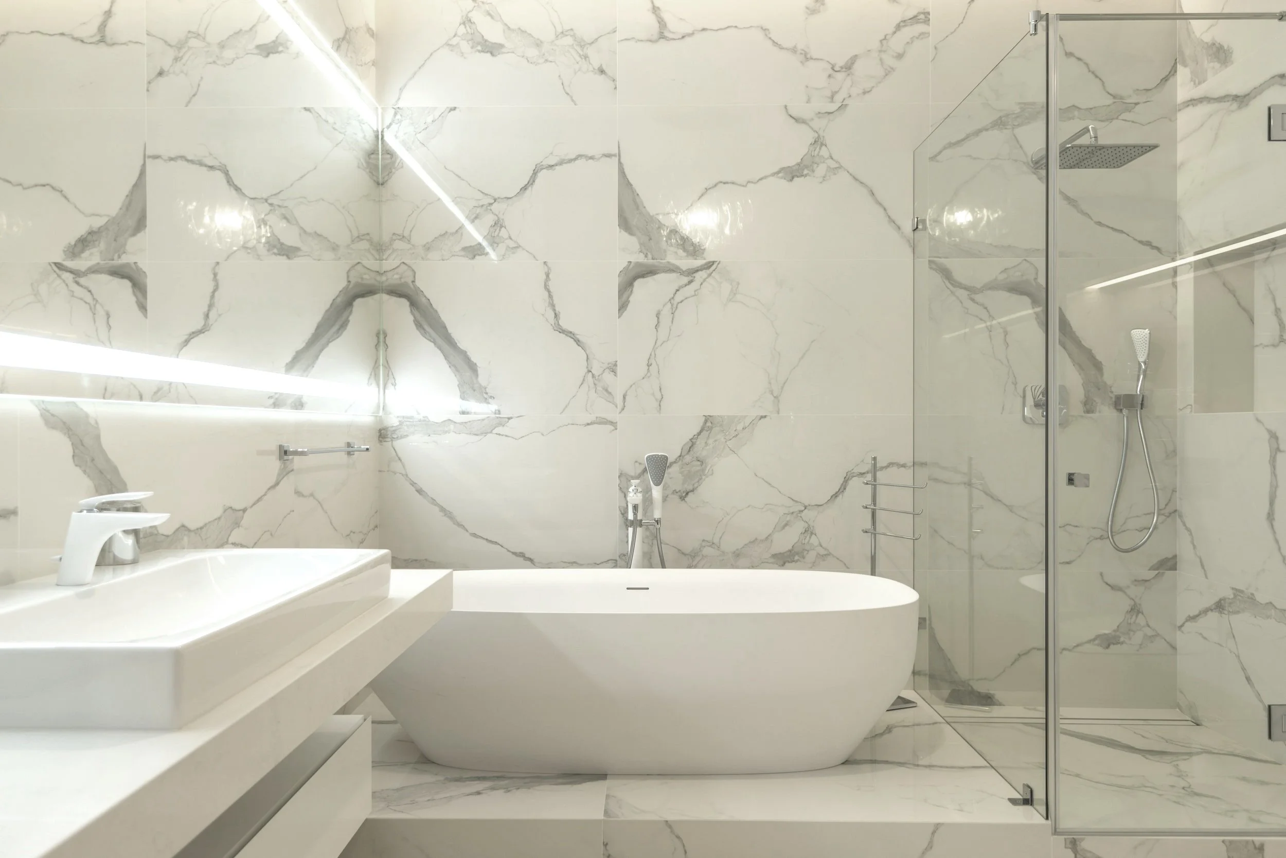 Modern bathroom with white marble walls and floors, featuring a free-standing bathtub, a glass-enclosed shower with rainfall and handheld showerheads, and a rectangular white sink with a minimalist faucet.