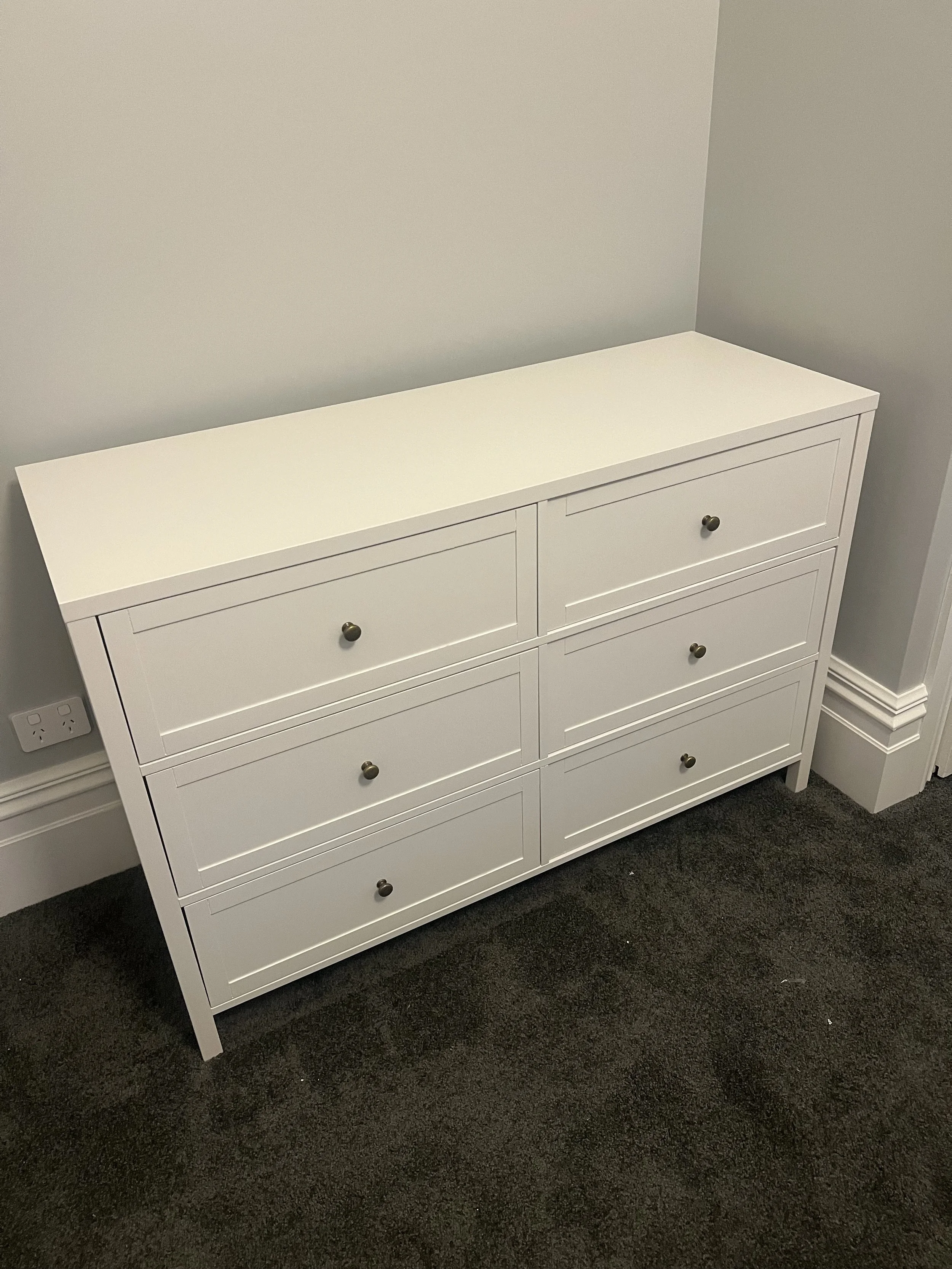White six-drawer dresser with small round knobs, placed against a light-colored wall and carpeted floor.