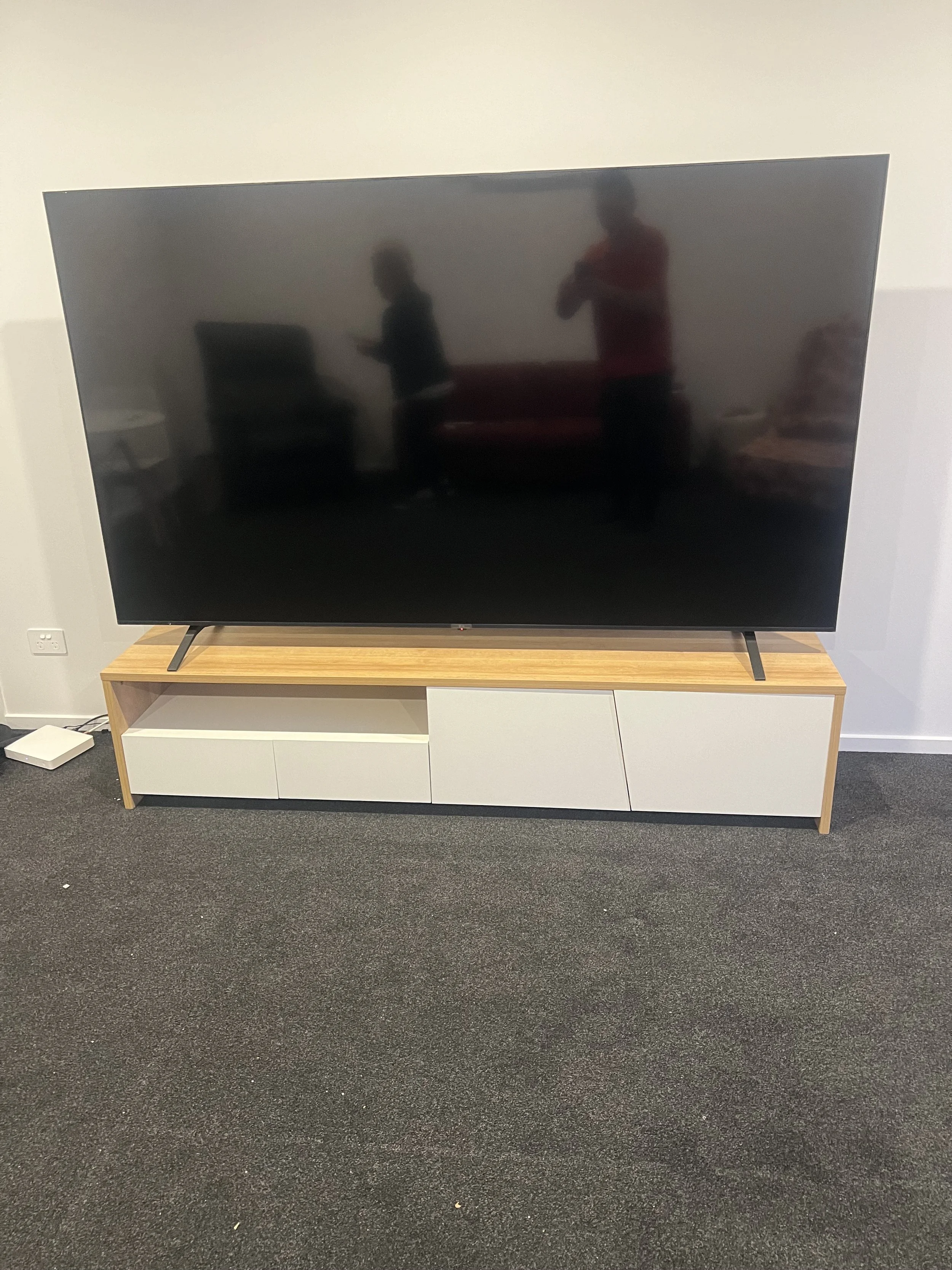 Large flat-screen television on a wooden TV stand with white drawers in a room with dark carpet and a white wall.