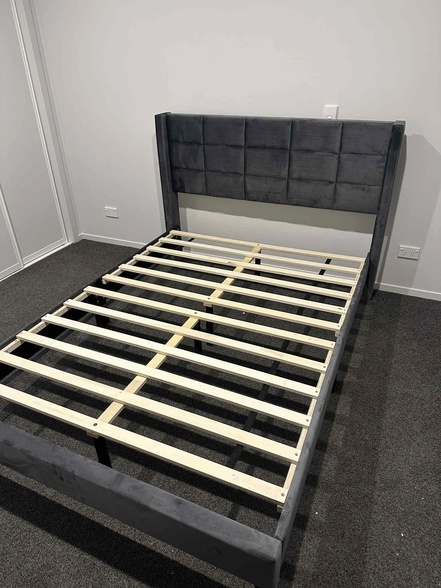 Unmade bed with a gray upholstered headboard and wooden bed frame on dark carpet in a room with white walls and electrical outlets.