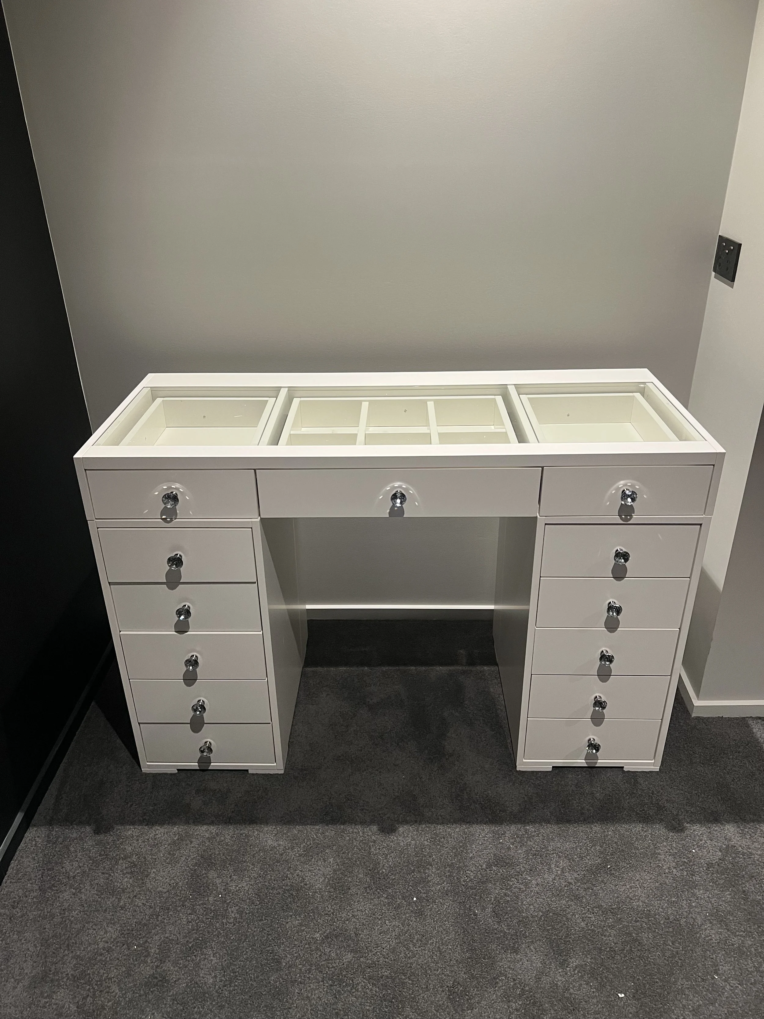 White jewelry display cabinet with glass top and multiple drawers, placed against a gray wall on a dark carpeted floor.