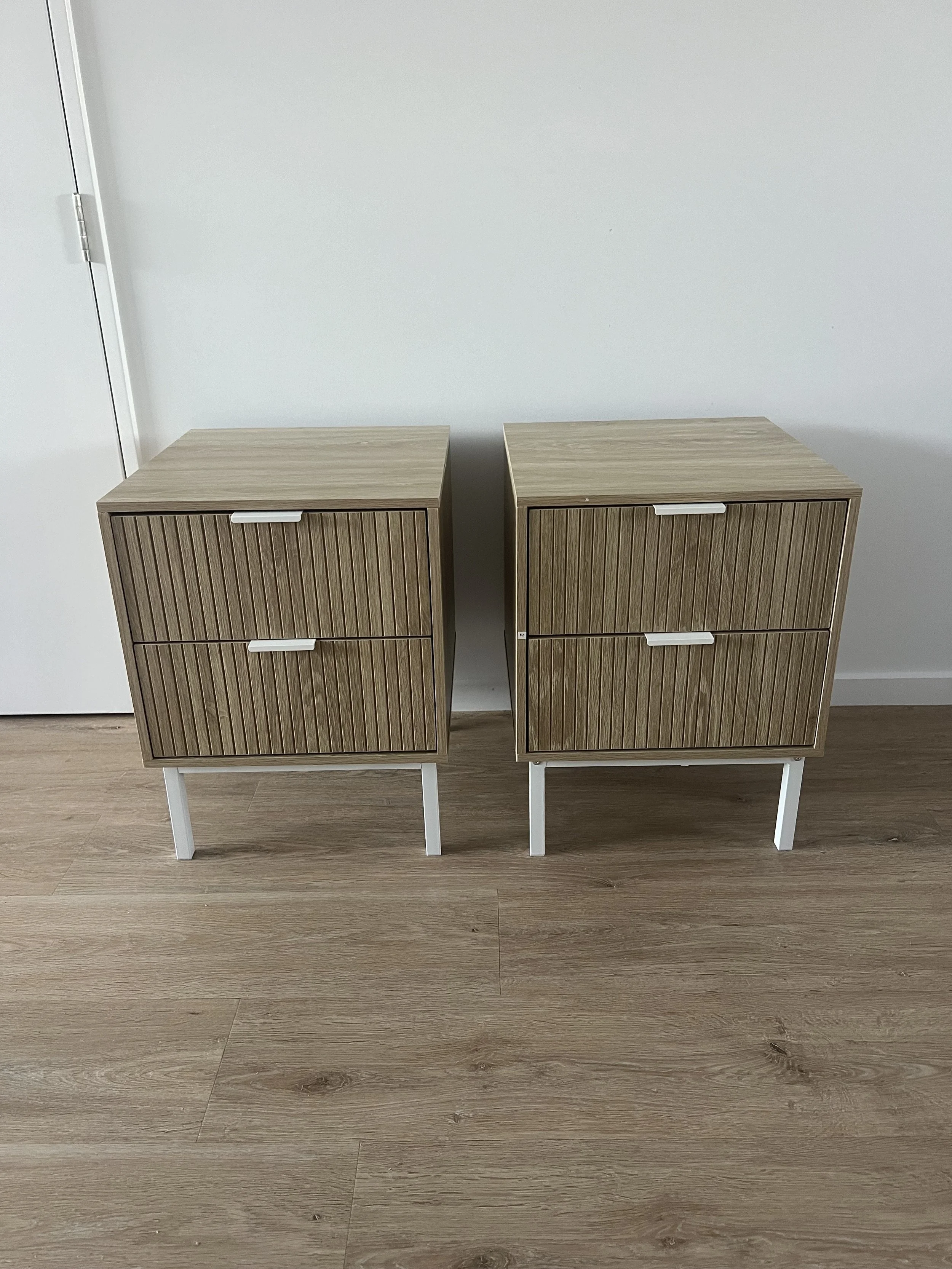 Two wooden nightstands with white legs and two drawers each, placed side by side against a white wall floor.