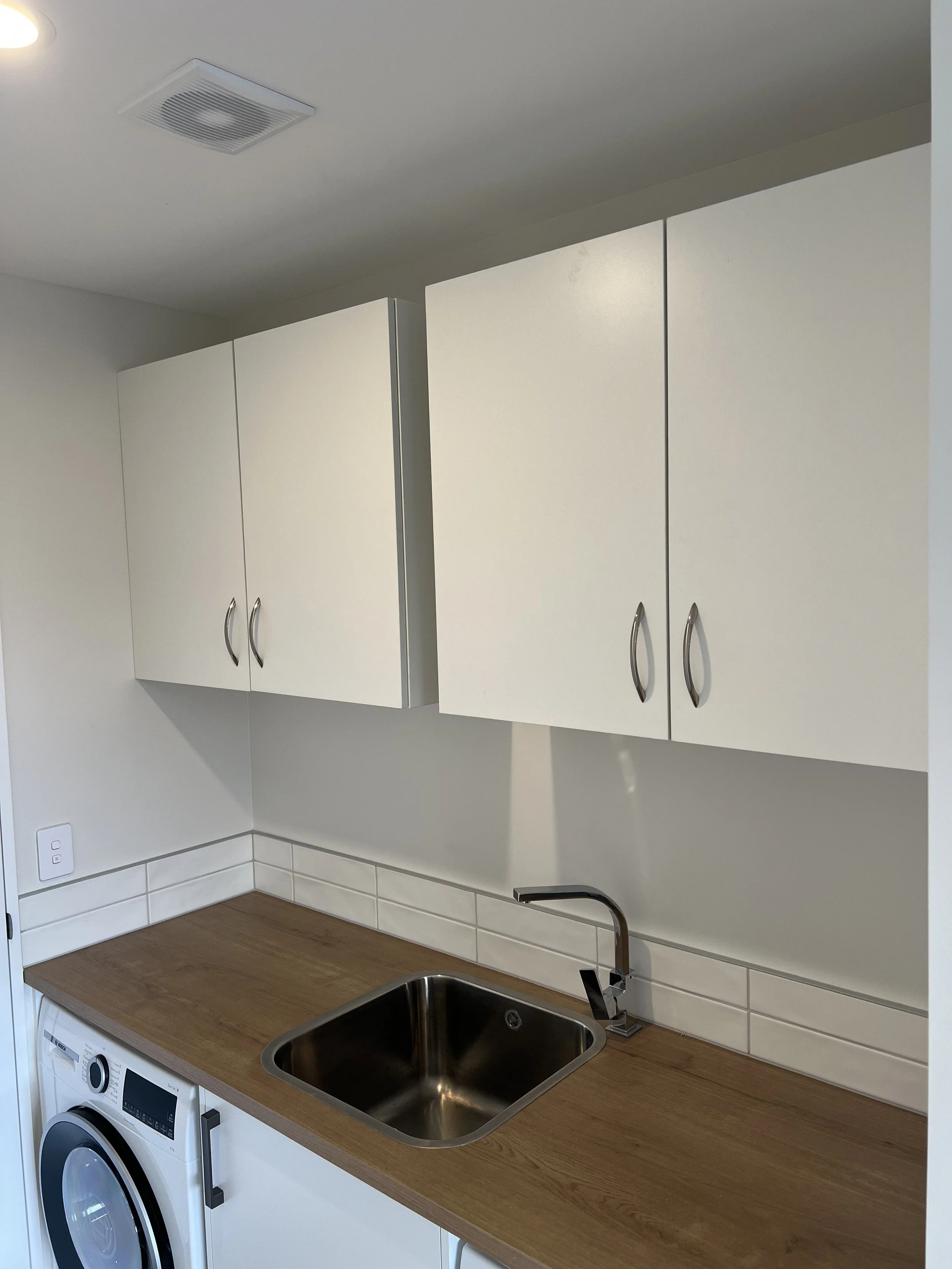 Kitchen corner with a wooden countertop, a stainless steel sink, white cabinets above, a white tiled backsplash, a washing machine underneath, and a ceiling vent.