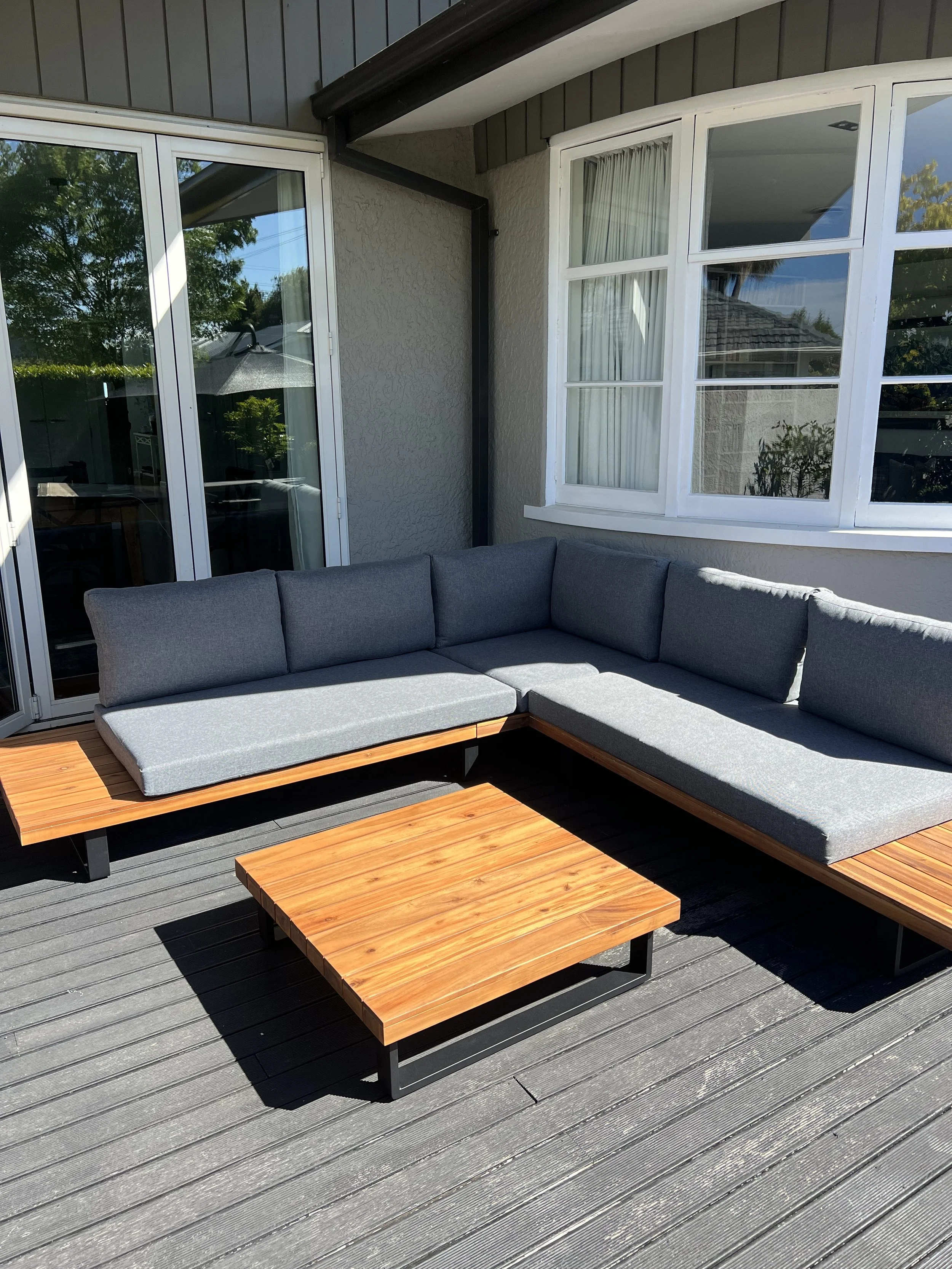 Outdoor patio with wooden deck, gray cushioned sectional sofa, and two matching wooden coffee tables, next to a house with sliding glass door and white-framed windows.