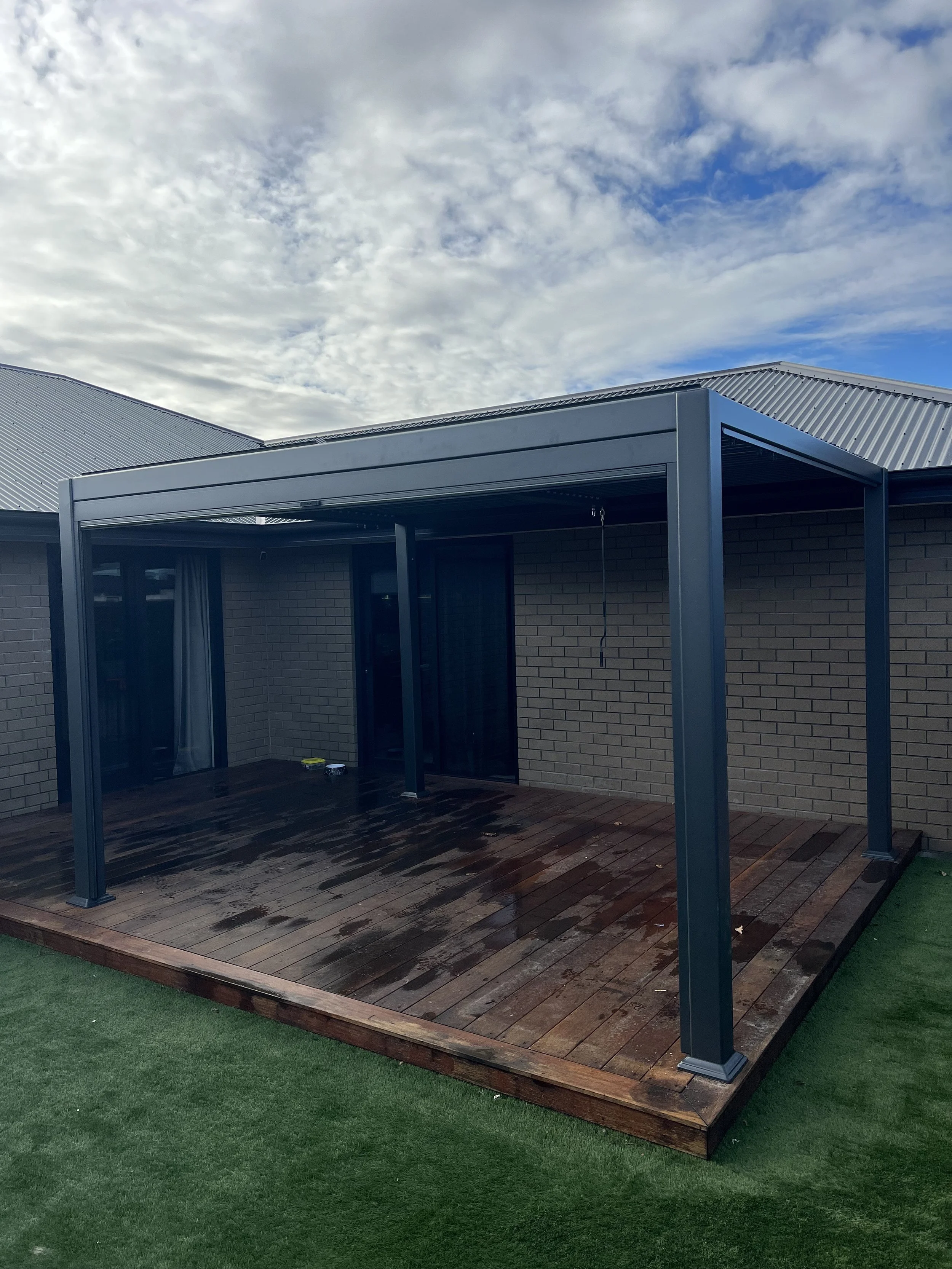 A wooden deck with wet surface, attached to a brick house with a metal roof, under a partly cloudy sky.
