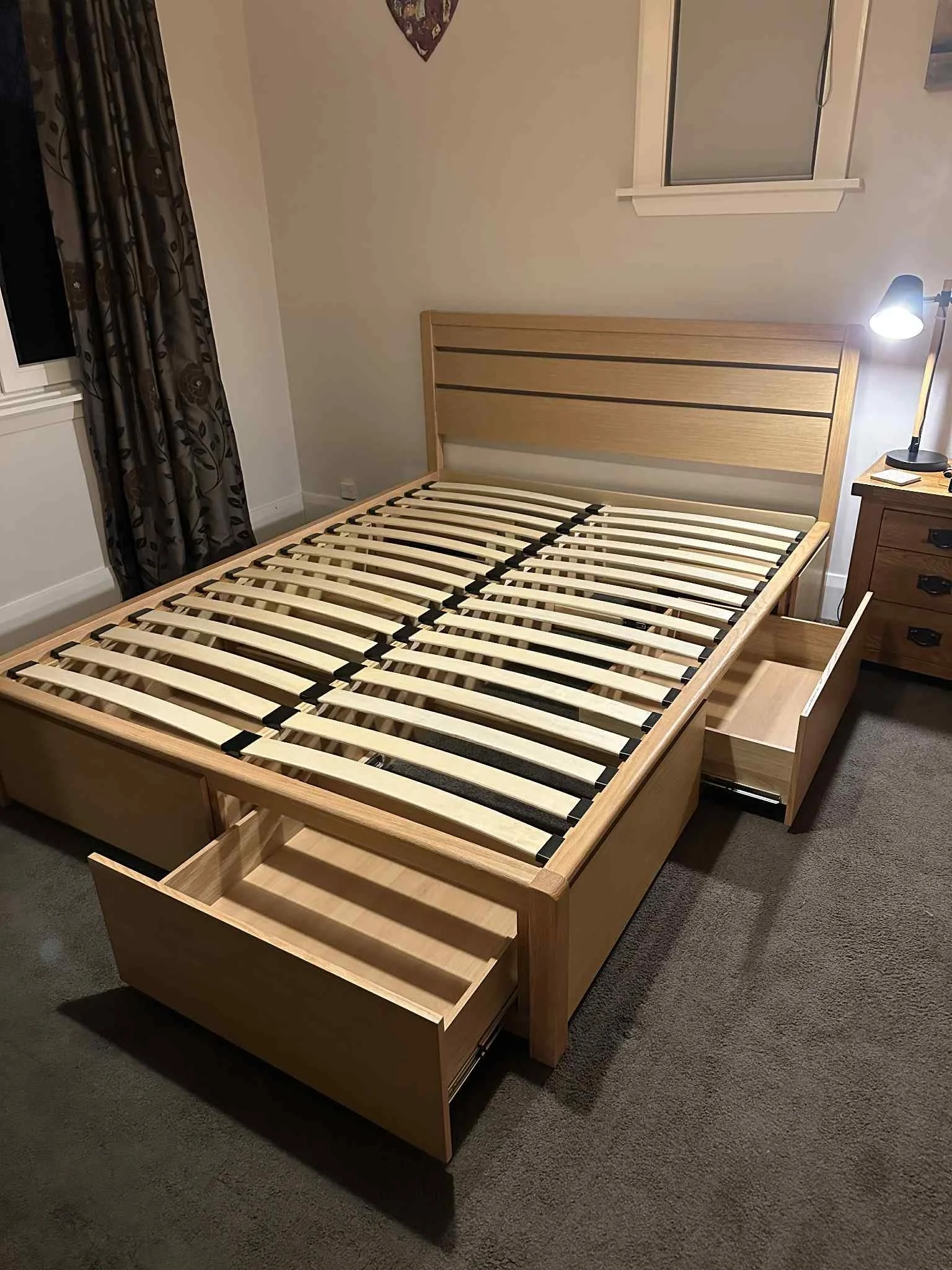 Unmade wooden bed frame with open drawers at the sides in a bedroom with a nightstand and a lamp