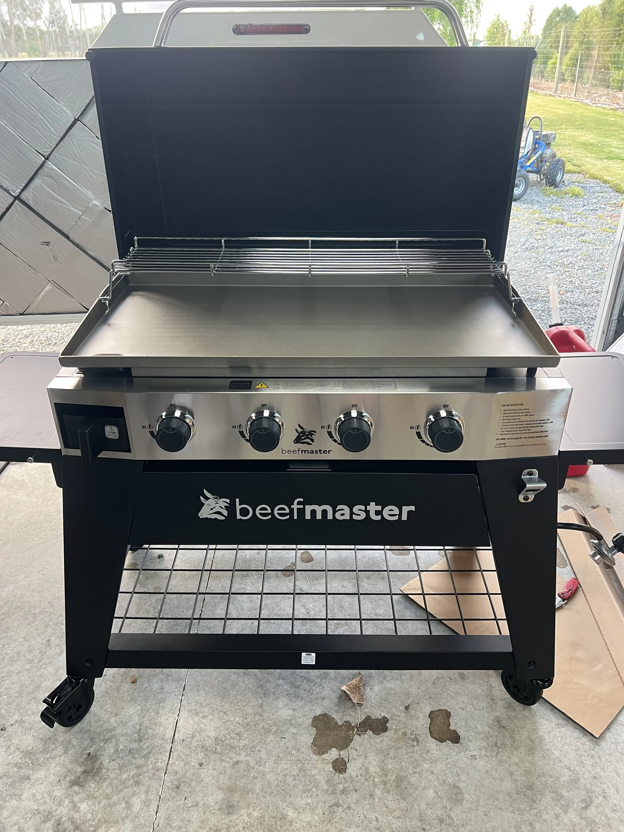 A large outdoor grill with the brand name 'beefmaster' on the front, four control knobs, and a metal table rack underneath. The grill is open, showing the cooking surface, and is situated on a concrete patio near some leaves and cardboard.