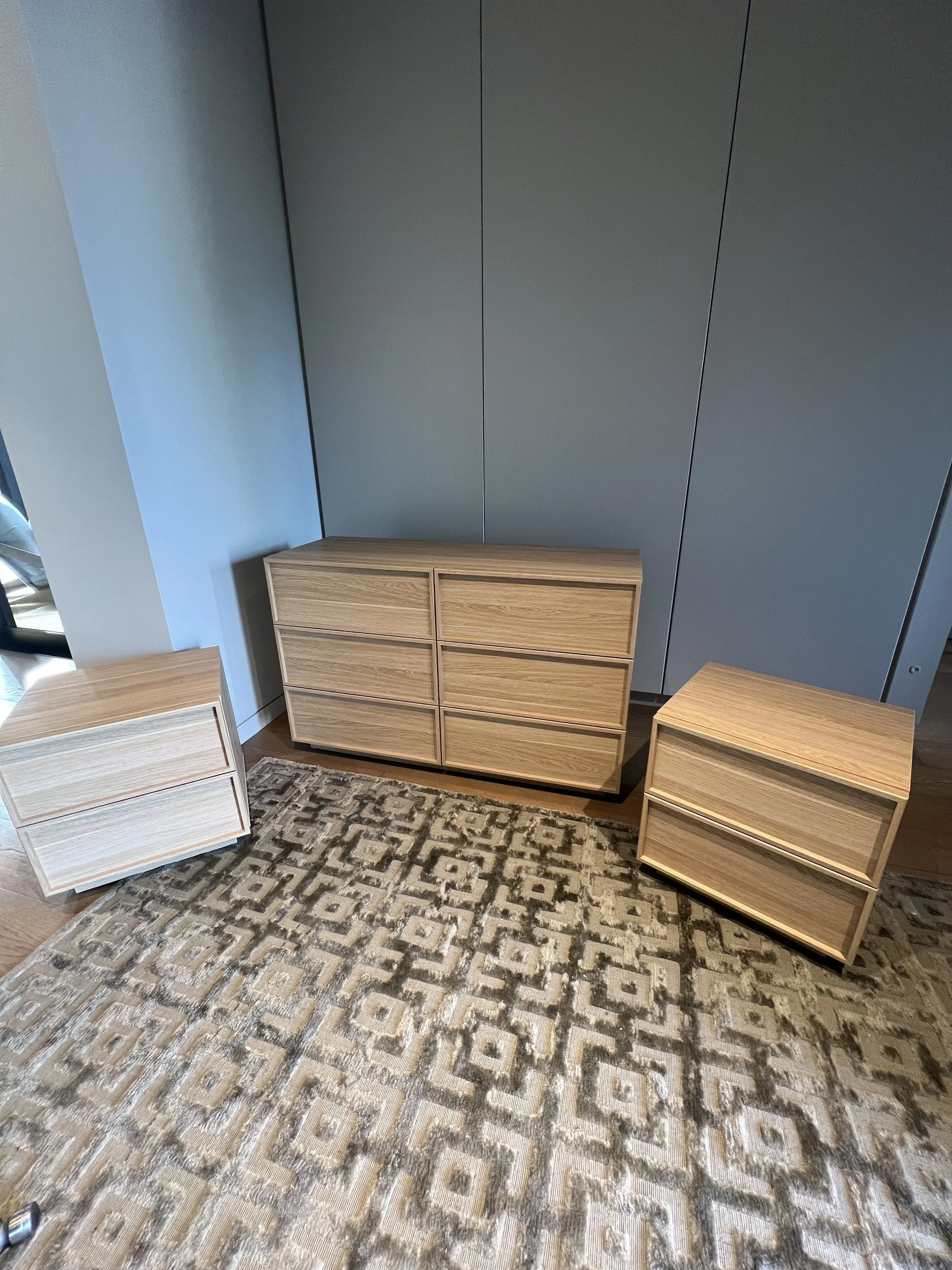 Three wooden dressers in a modern room with a patterned beige and brown rug and gray wall panels.