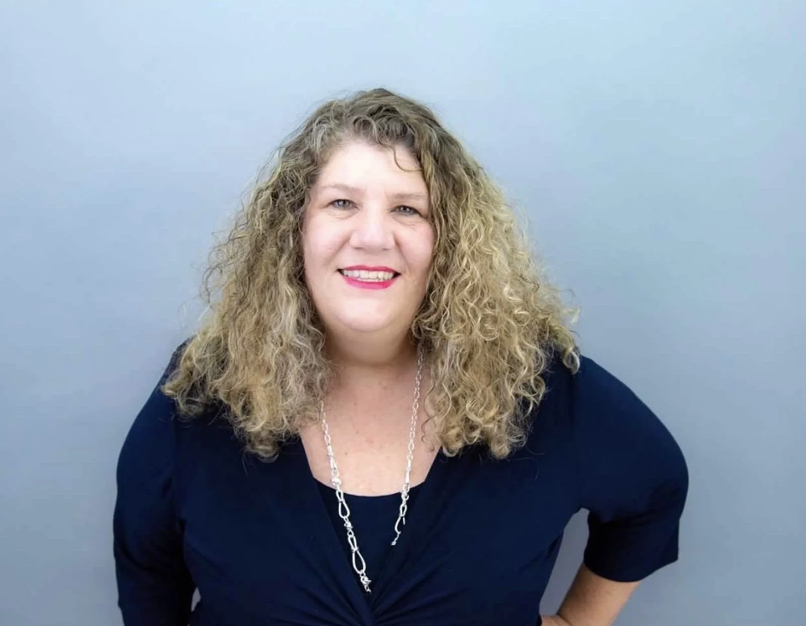 A woman with curly blonde hair smiling, wearing a black top and a silver chain necklace, against a plain light blue background.