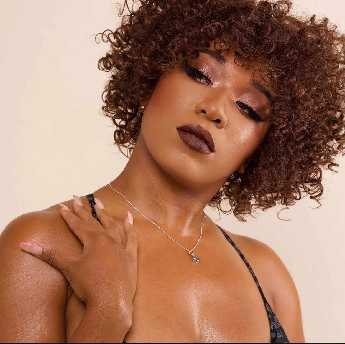 A woman with curly brown hair, wearing makeup with dark lipstick, wearing a thin necklace with a small pendant, and a black strap top. She is touching her shoulder with her left hand.