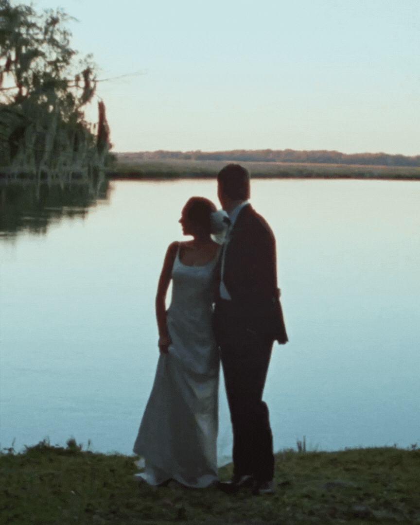 Silhouetted couple in formal attire standing by a lake at sunset.