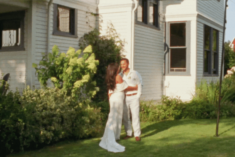 Black and white photo of a couple embracing outdoors, with trees and bushes in the background.