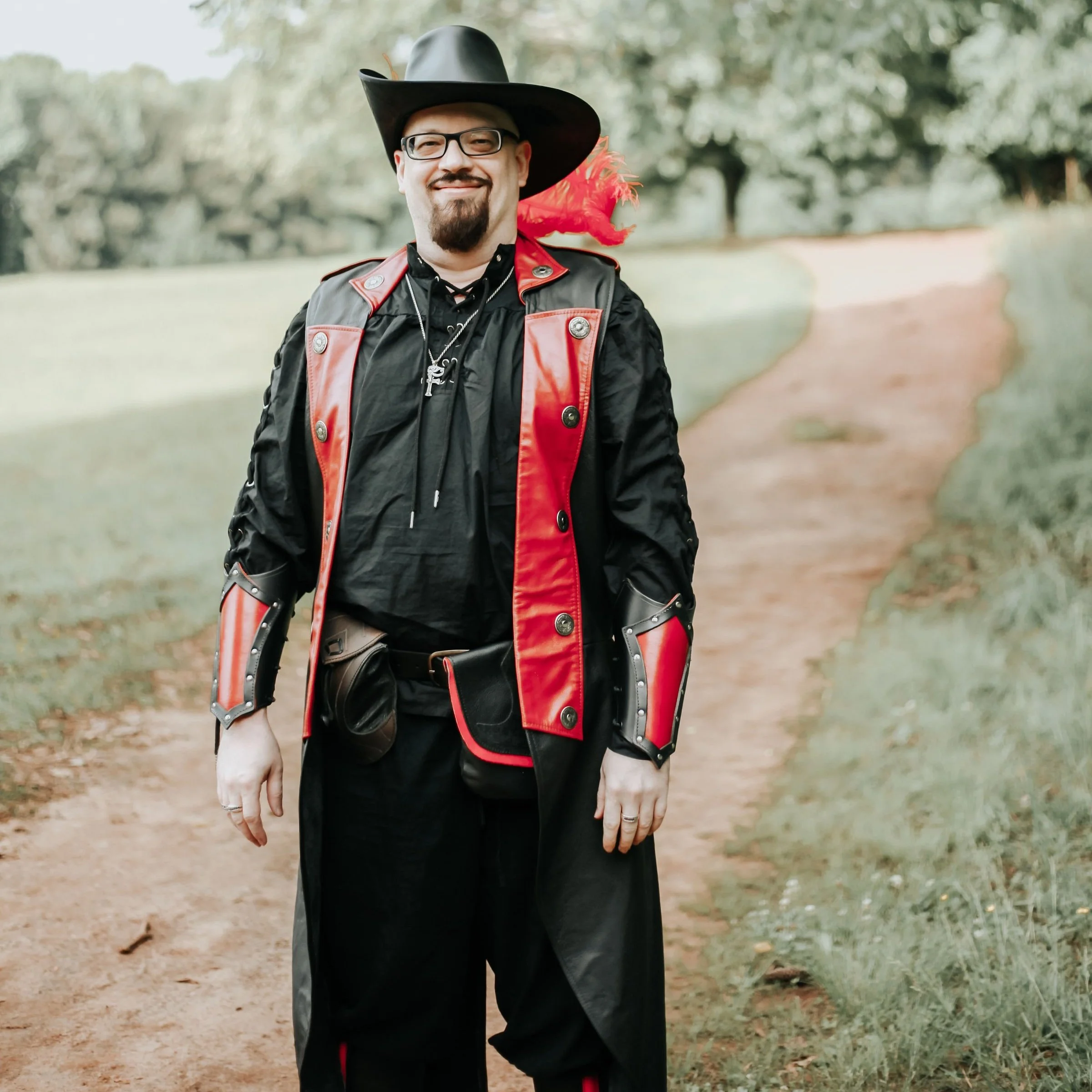 A man dressed in a fantasy or cosplay costume with black and red armor, a black wide-brimmed hat with a red feather, glasses, and bearded, standing on a dirt path in a park.