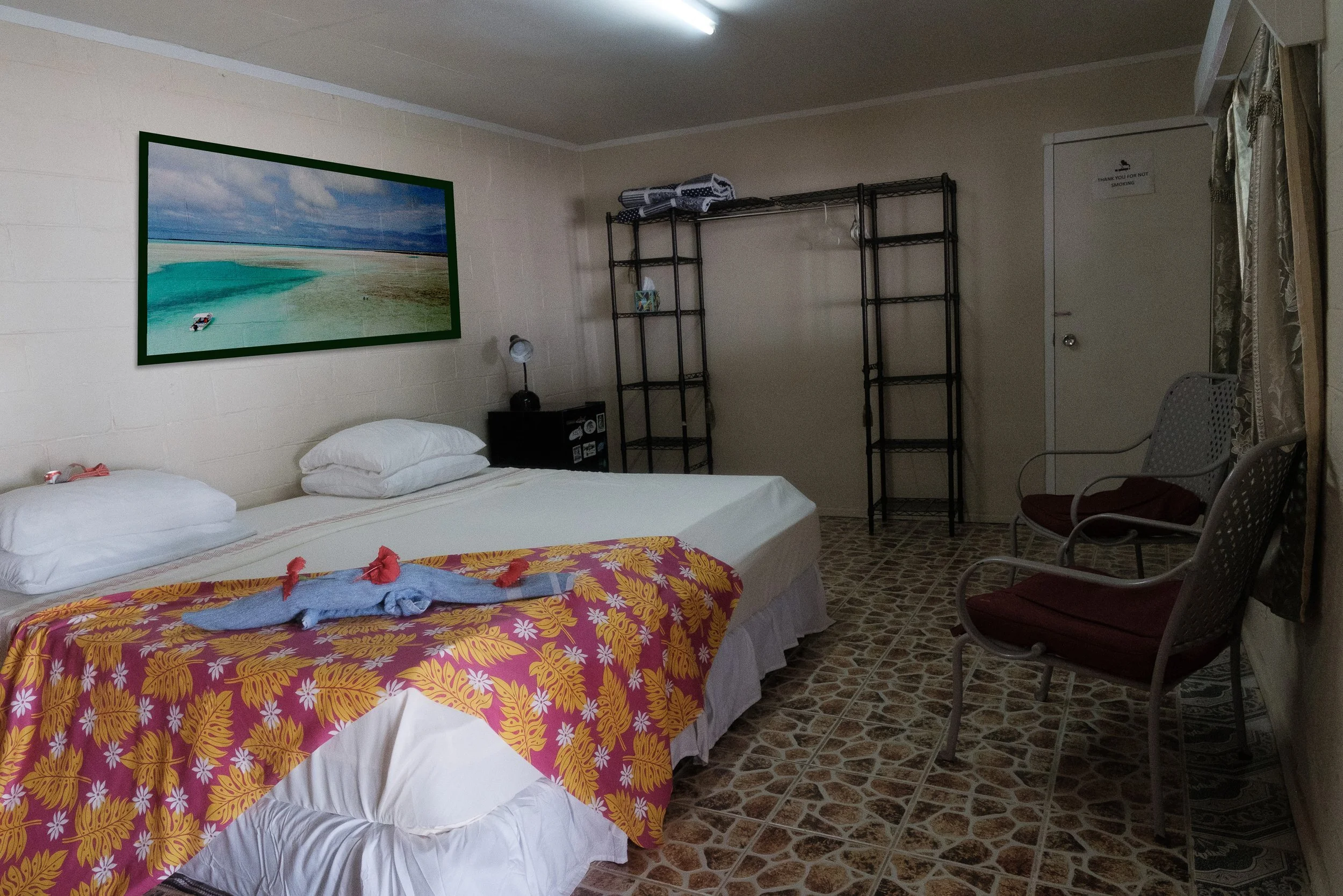 A hotel room with two beds, a colorful bedspread, a towel animal on the bed, a painting of a beach scene, a small nightstand, and a black shelving unit.