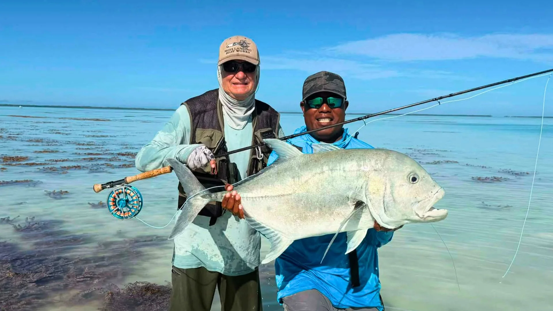 Man holding a large fish near the shore with a fishing rod on his back under partly cloudy sky.