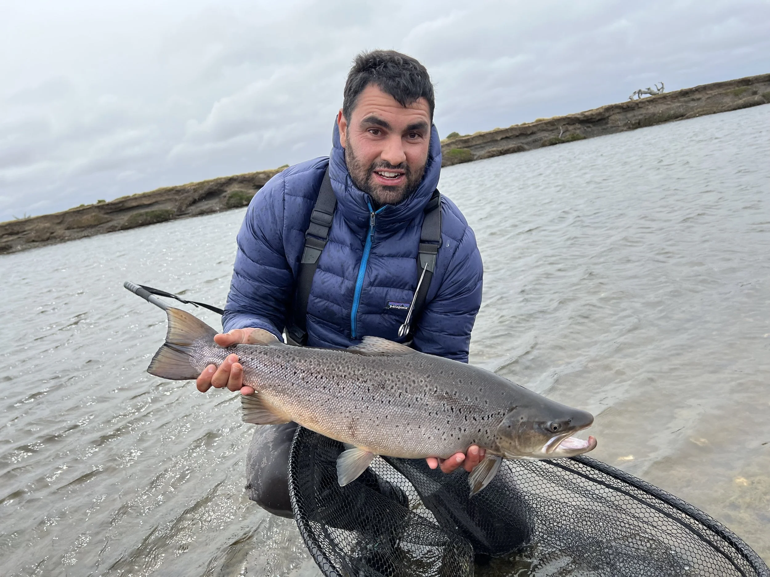 Man kneeling in water holding a large fish, wearing a blue jacket with a backpack, in a cloudy outdoor setting