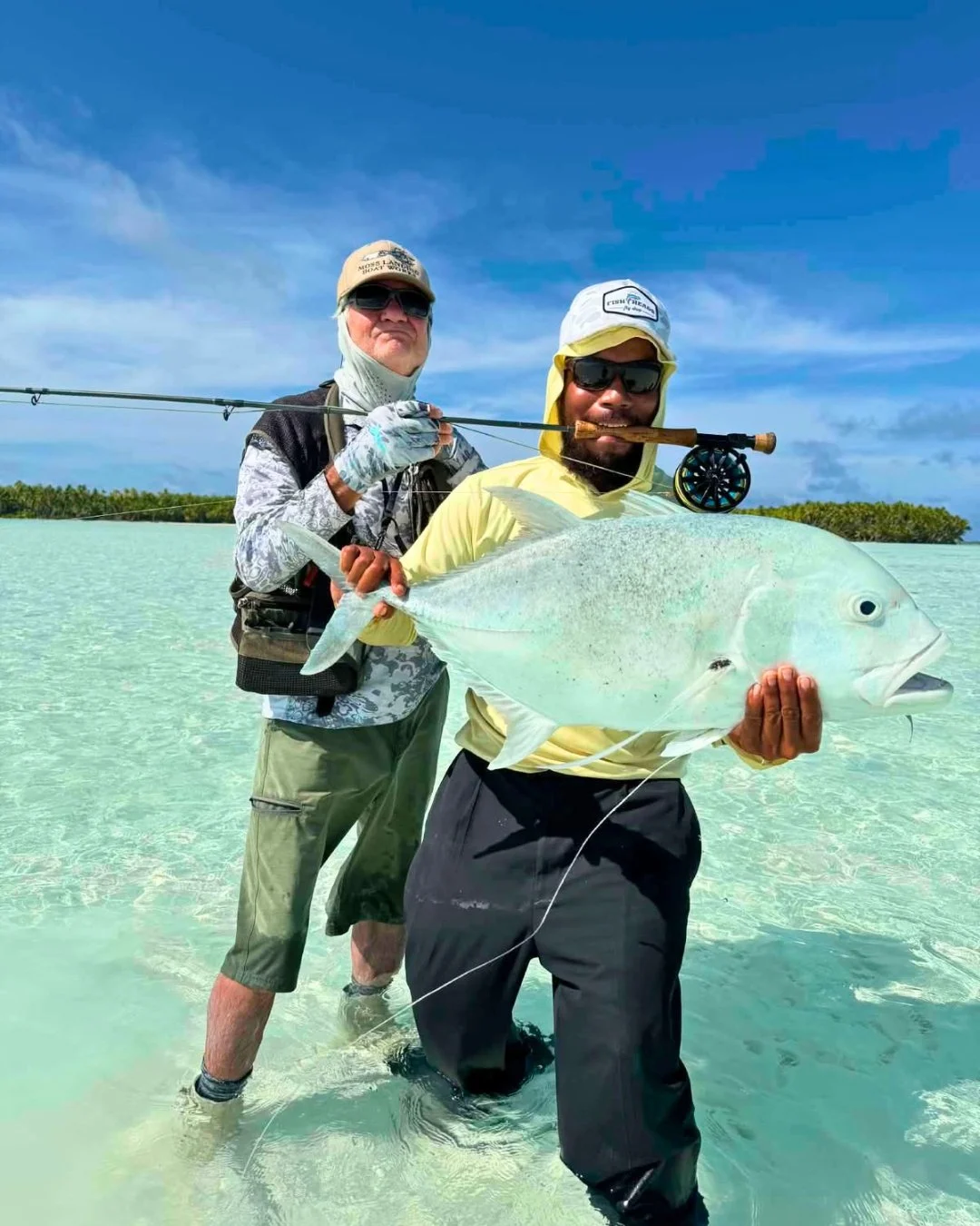 Man holding a large fish in shallow water, with a fishing rod on his shoulder and a clear sky in the background
