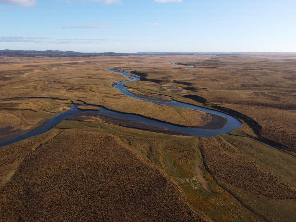 Aerial view of a winding river flowing through a flat, open landscape with patches of grass and fields under a partly cloudy sky.