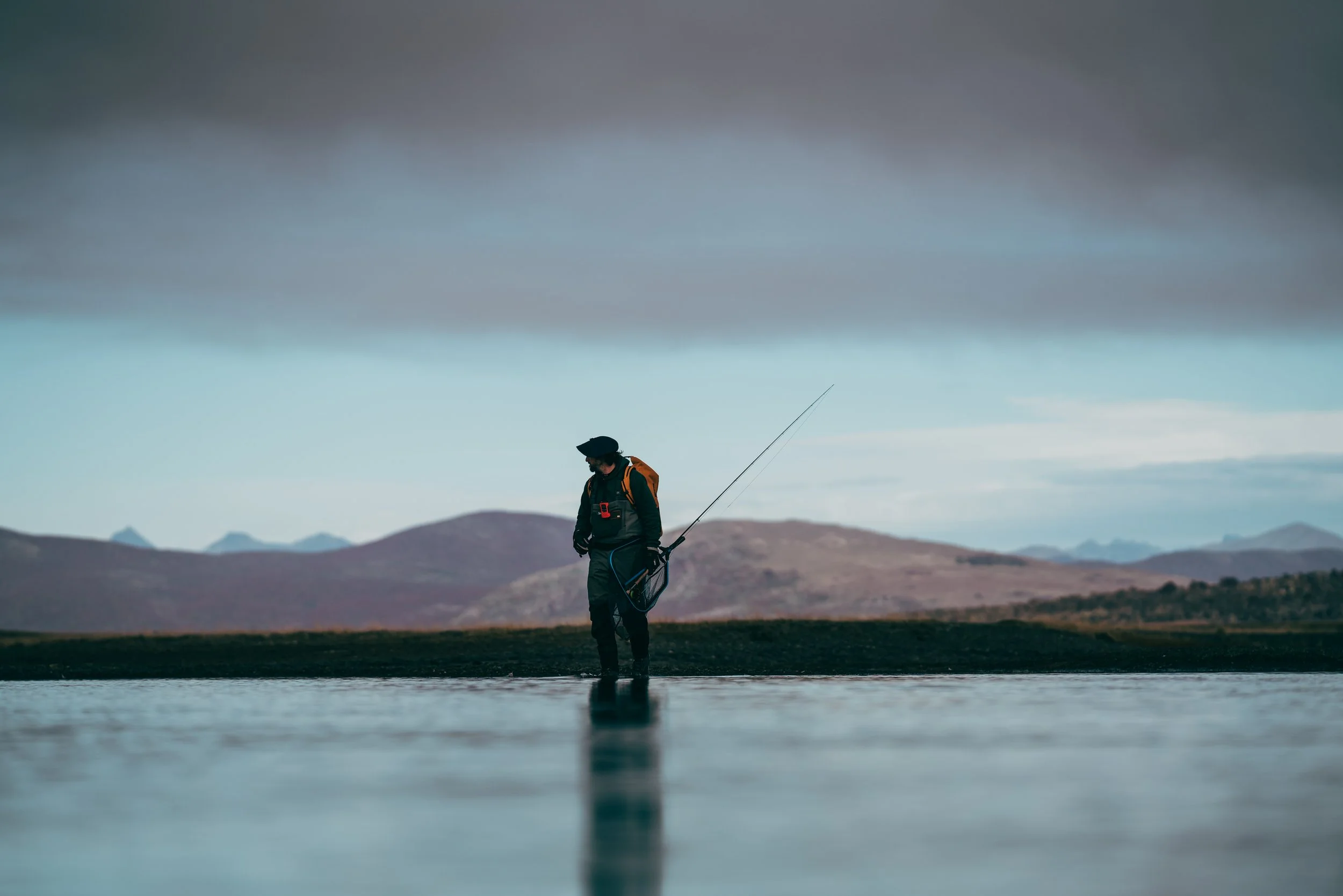 Person standing in shallow water holding a fishing rod with mountains and cloudy sky in the background.