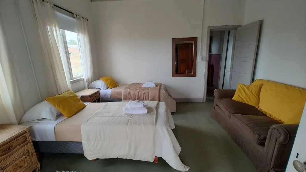 A bedroom with two beds, each with white sheets and yellow pillows, a brown couch with yellow pillows, and windows with white curtains.
