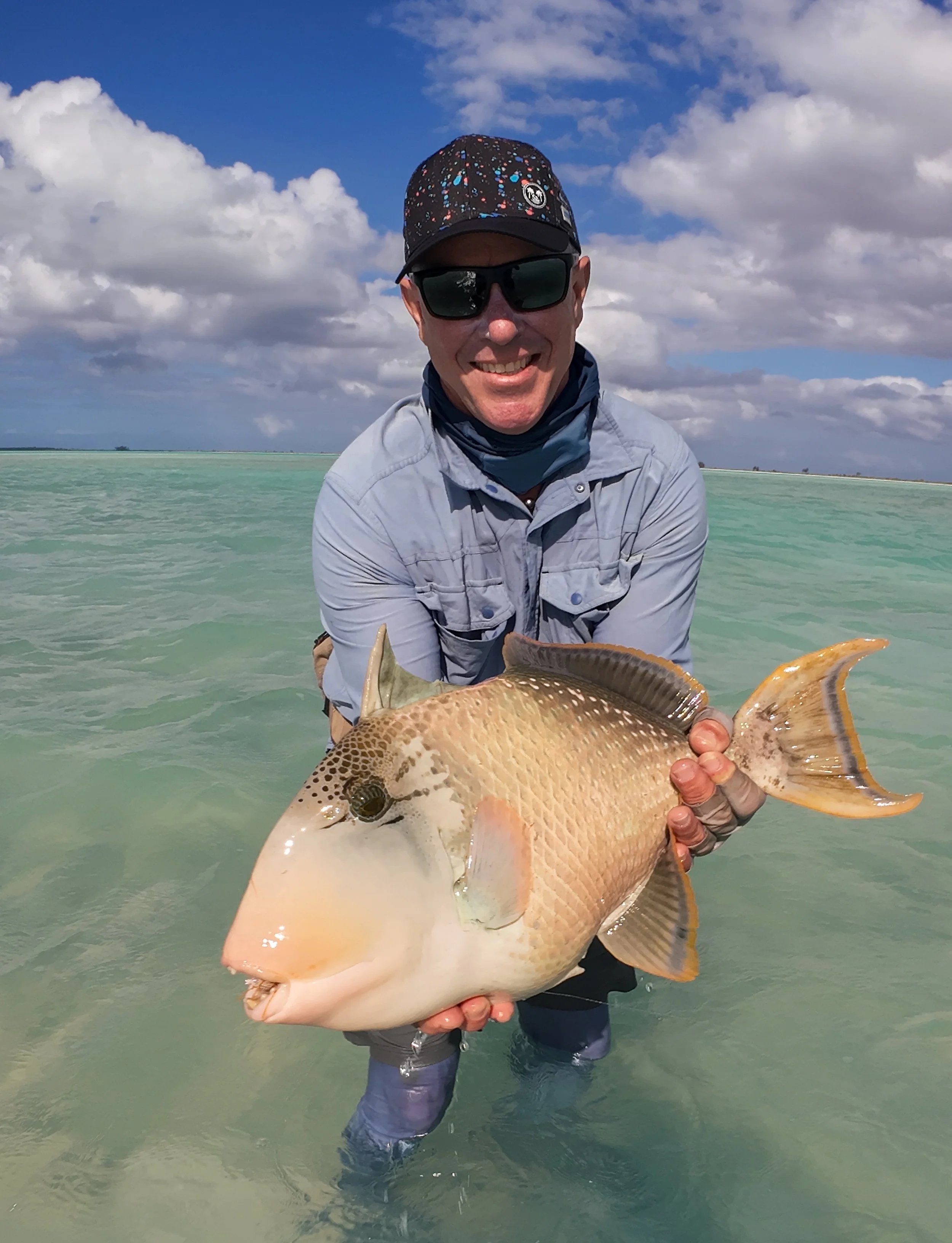 A man wearing sunglasses, a hat, and fishing gear standing in shallow ocean water holding a large fish with a light-colored body and dark markings near its face and tail.