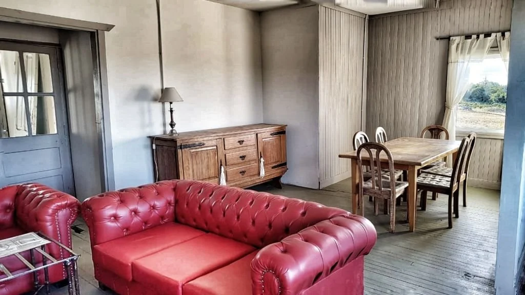 Living room with a red tufted sofa, wooden sideboard with lamp, and a dining table with six chairs near a window showing an outdoor landscape.