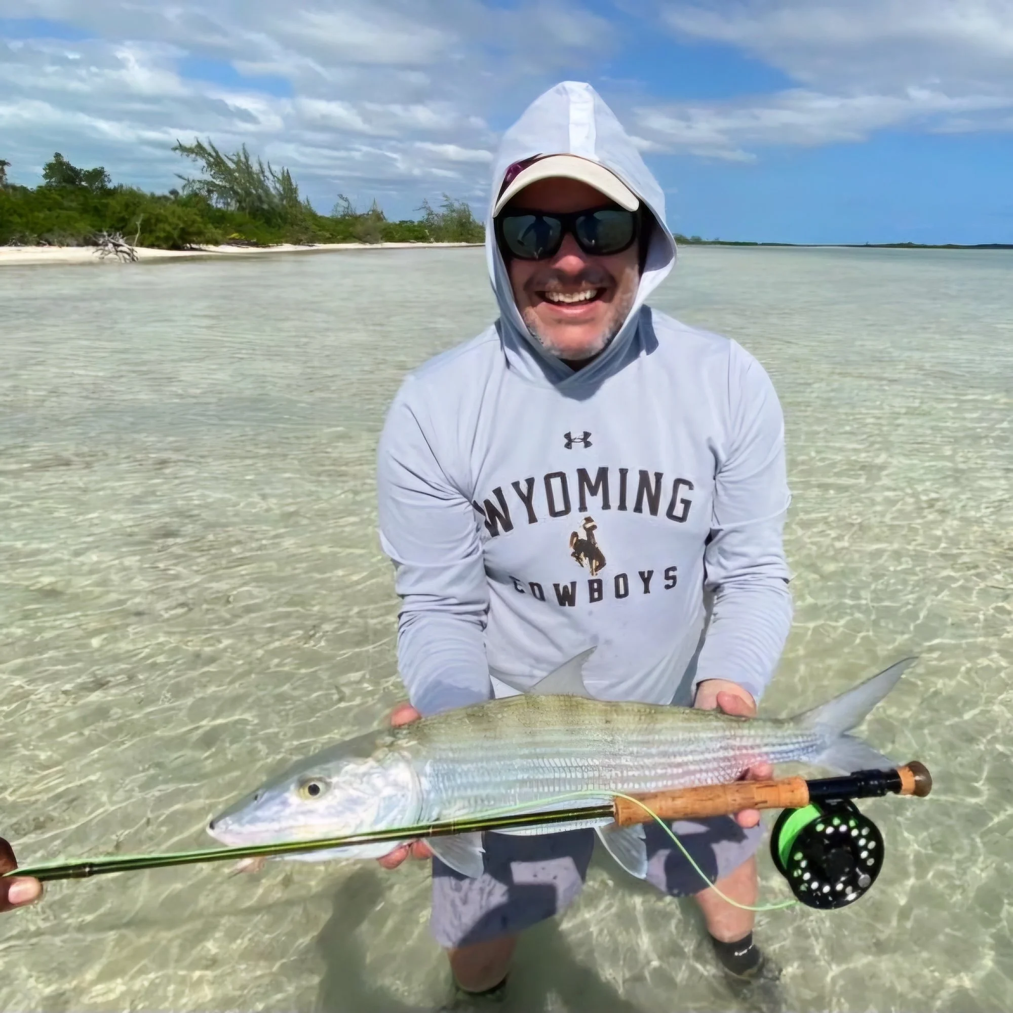Man in white hoodie and sunglasses holding a large fish on a fishing line in shallow water near a sandy shoreline with bushes and cloudy sky in the background.