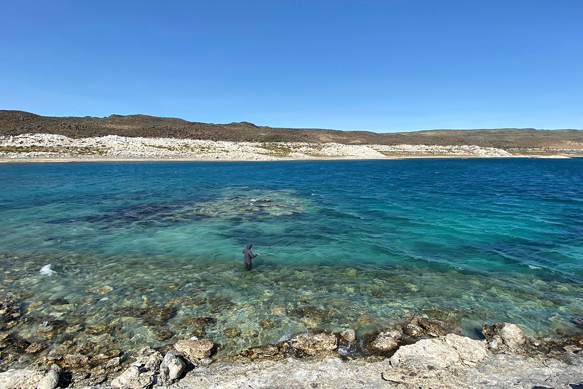 A person fishing in a clear, blue lake with a rocky shoreline and rolling hills in the background under a bright, clear sky.