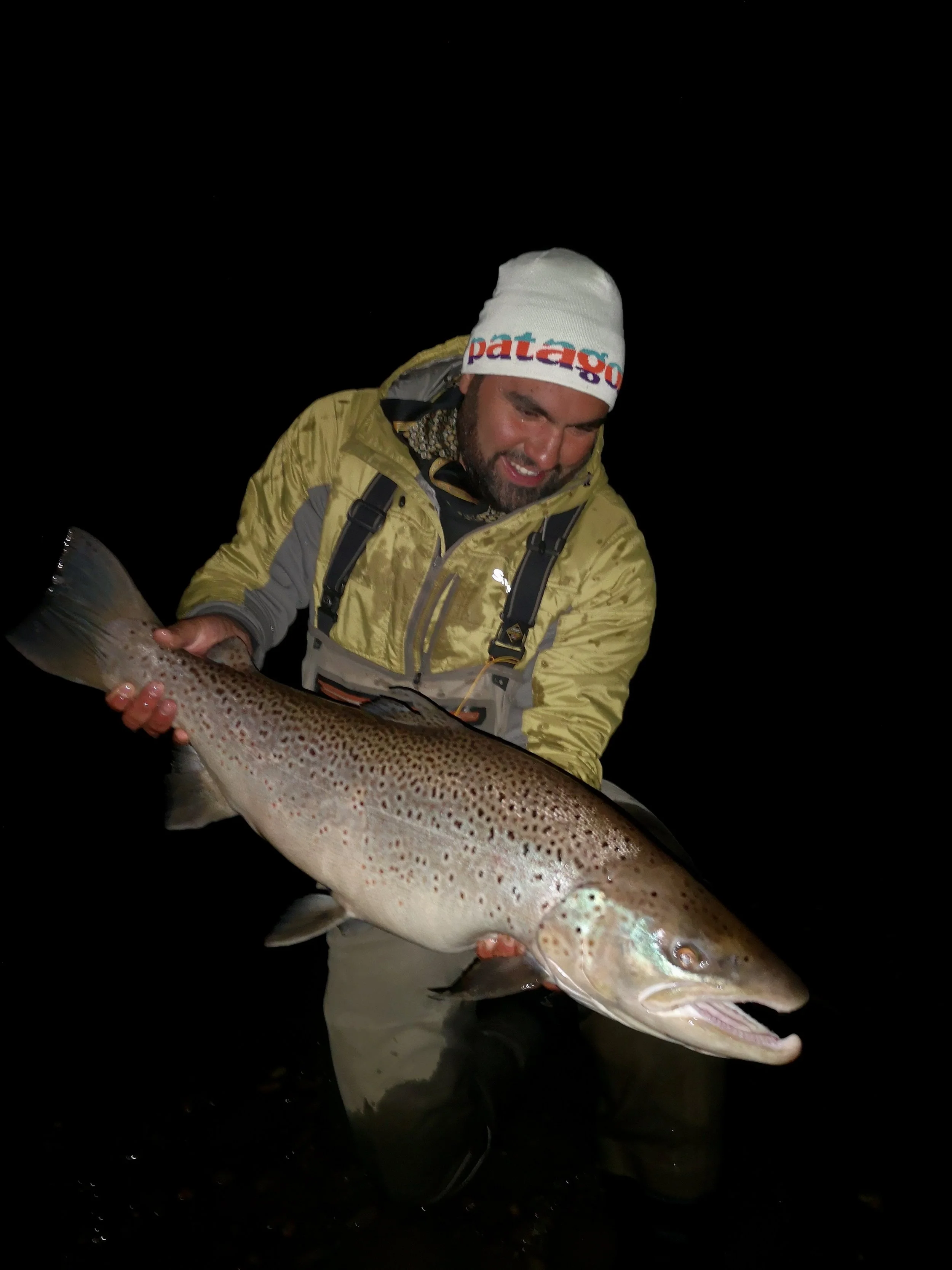 Man in yellow jacket holding a large rainbow trout at night, smiling and looking down at the fish, dark background.