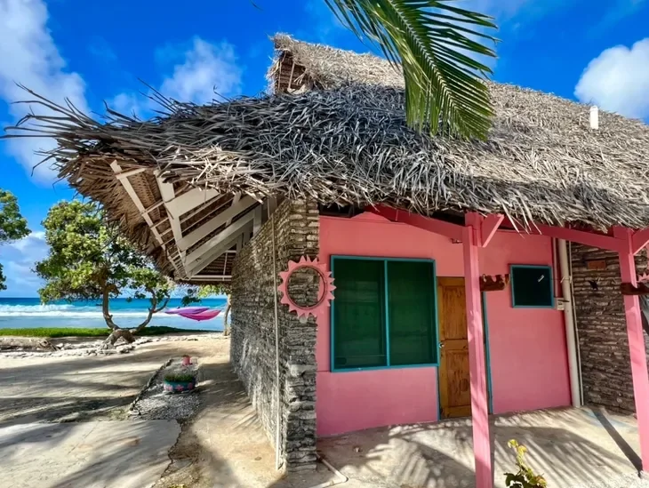A tropical beach house with pink walls, a thatched roof, and a palm tree in the foreground. The background shows sandy beach, ocean, and partly cloudy sky.