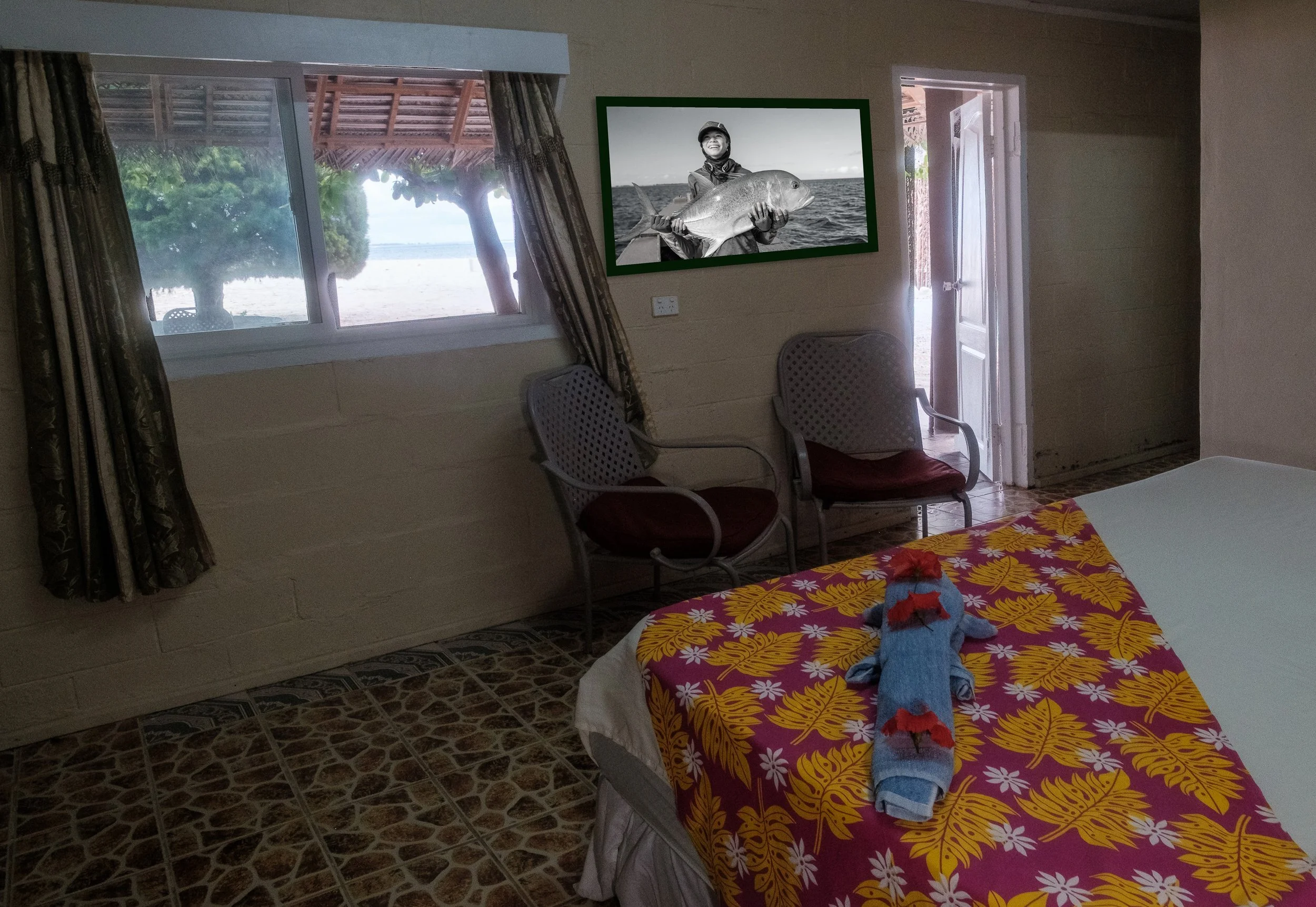 A bedroom with a bed covered in a vibrant yellow and red floral blanket and a stuffed animal resembling a lion on top. There are two chairs with maroon cushions near the wall, a window with patterned curtains showing trees outside, and a door leading outside. A framed black and white photo of a person holding a large fish hangs on the wall.