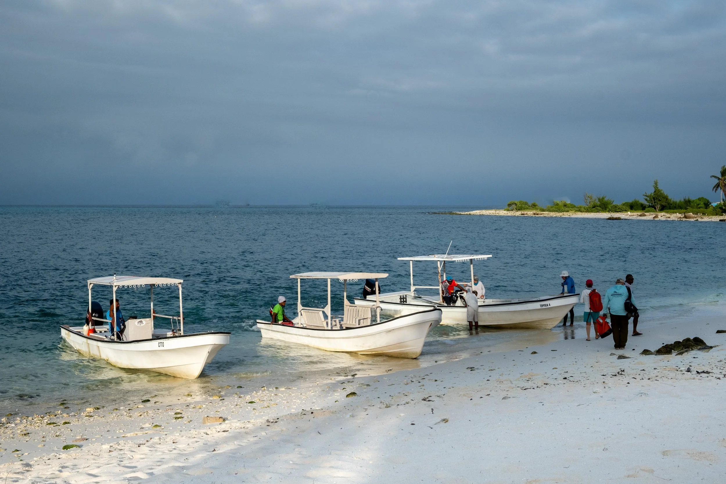 Several small white boats are on a sandy beach, with some people preparing to take them out on the water. The beach has white sand and a few rocks, with a backdrop of calm blue ocean and a cloudy sky.