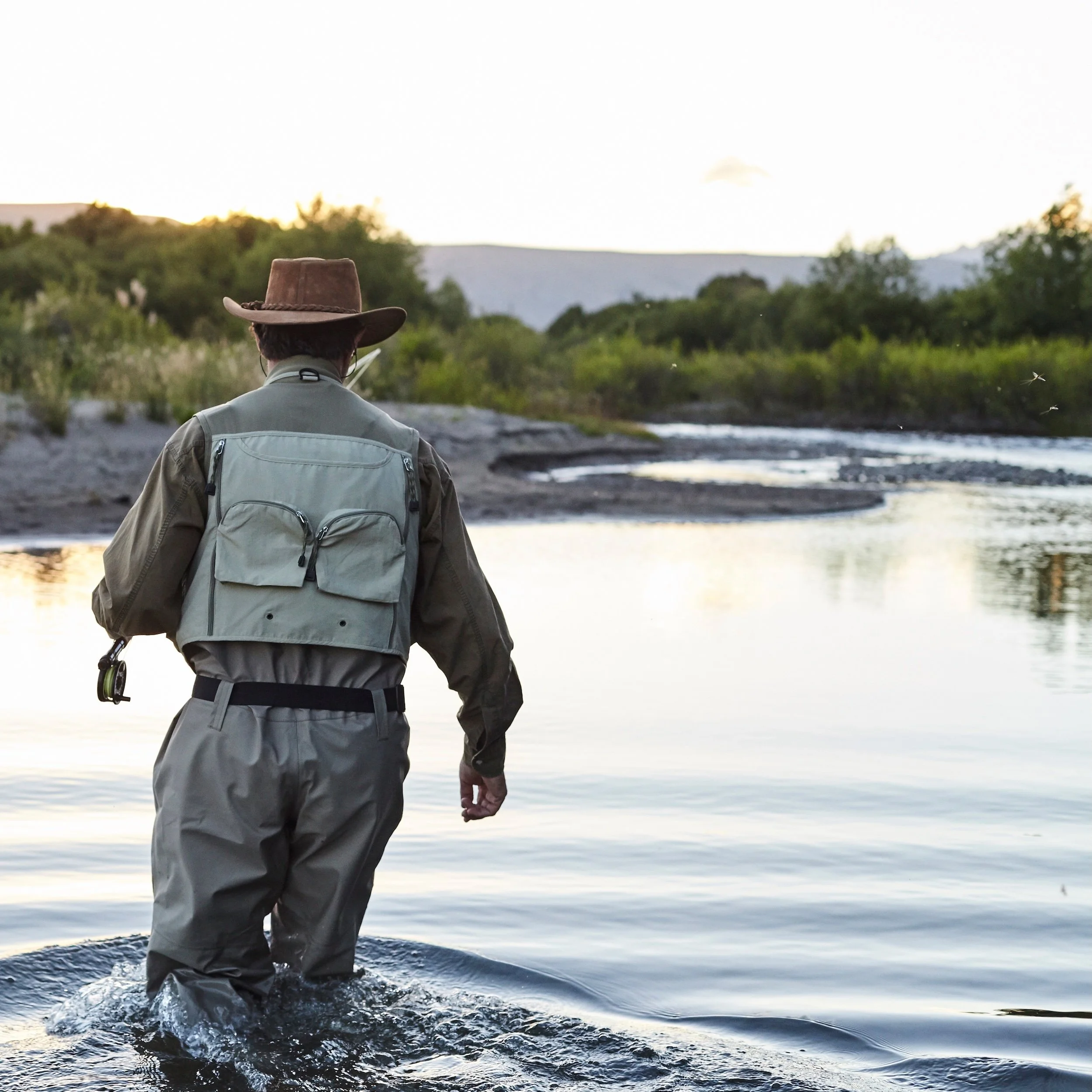 A person wearing a wide-brimmed hat, outdoor gear, and a backpack wading through a river at dusk, surrounded by green trees and distant mountains.